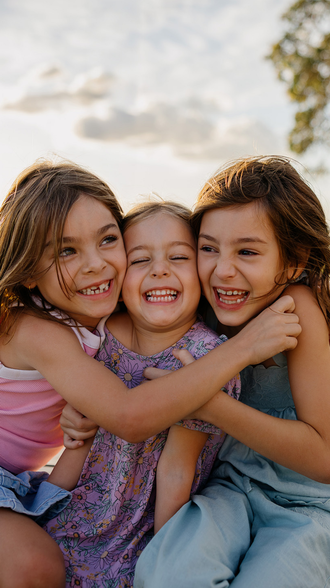 Three young girls sit close together outdoors, smiling and hugging each other, with a white fence, grass, and water in the background on a sunny day.