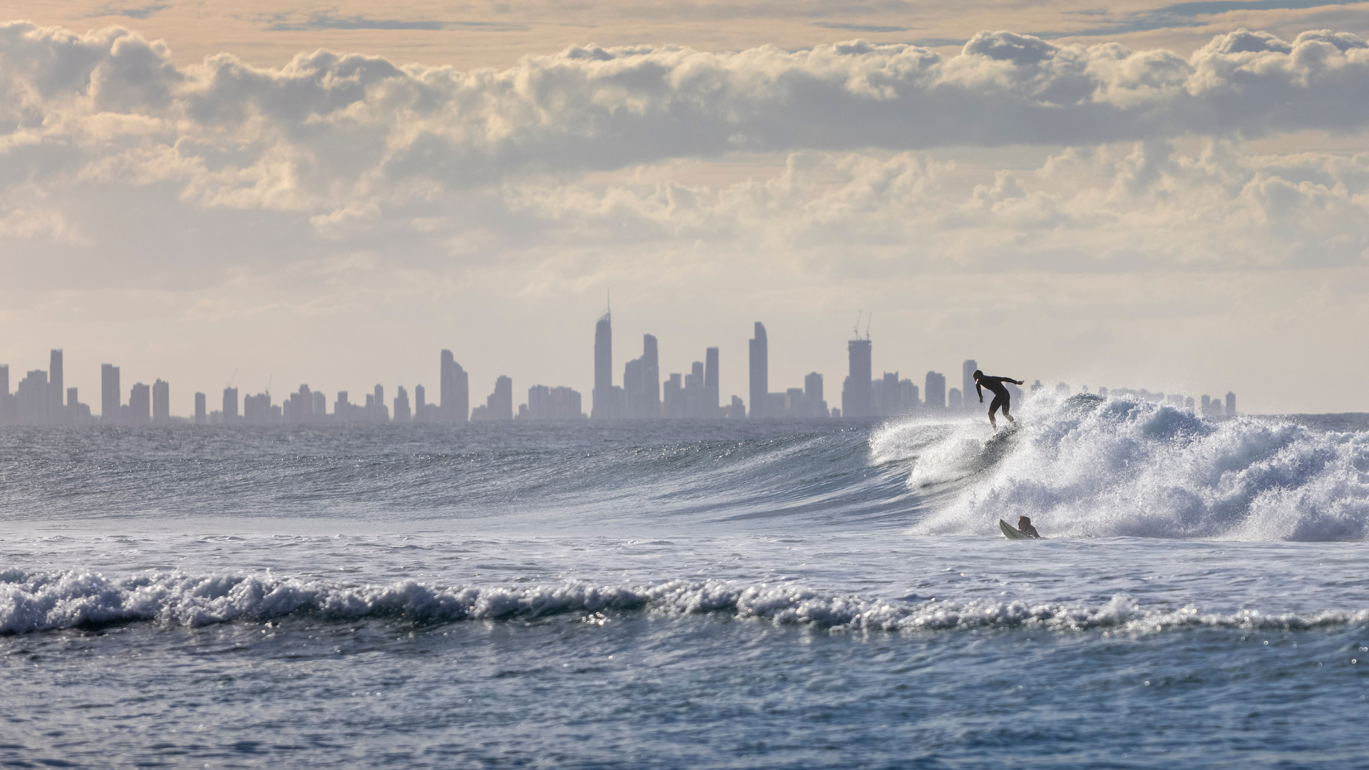 Surfer riding a wave in the ocean with Gold Coast city skyline in the background under a cloudy sky.