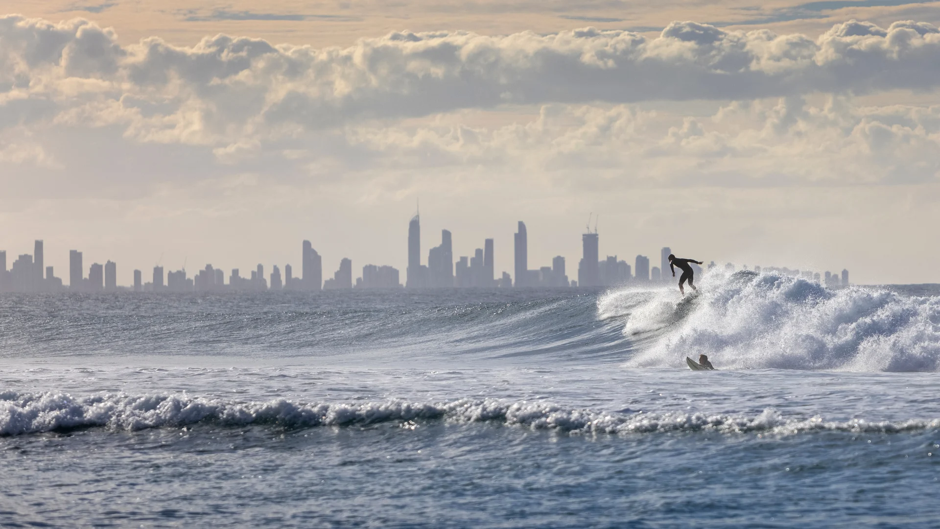 Surfer riding a wave in the ocean with Gold Coast city skyline in the background under a cloudy sky.