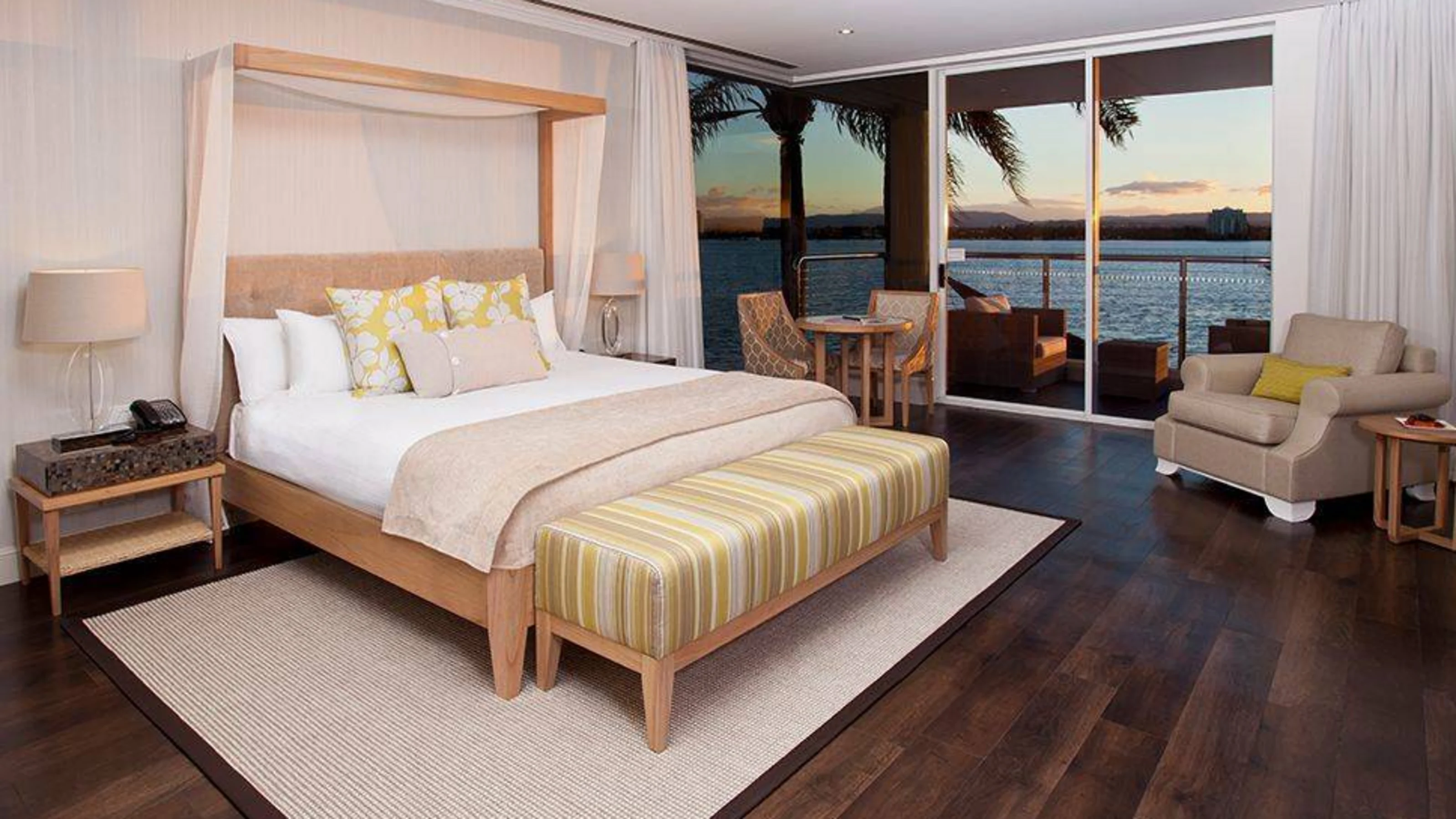 Hotel room with large bed, striped ottoman, armchair, and small dining area. Sliding glass doors open to a balcony overlooking a body of water at sunset.