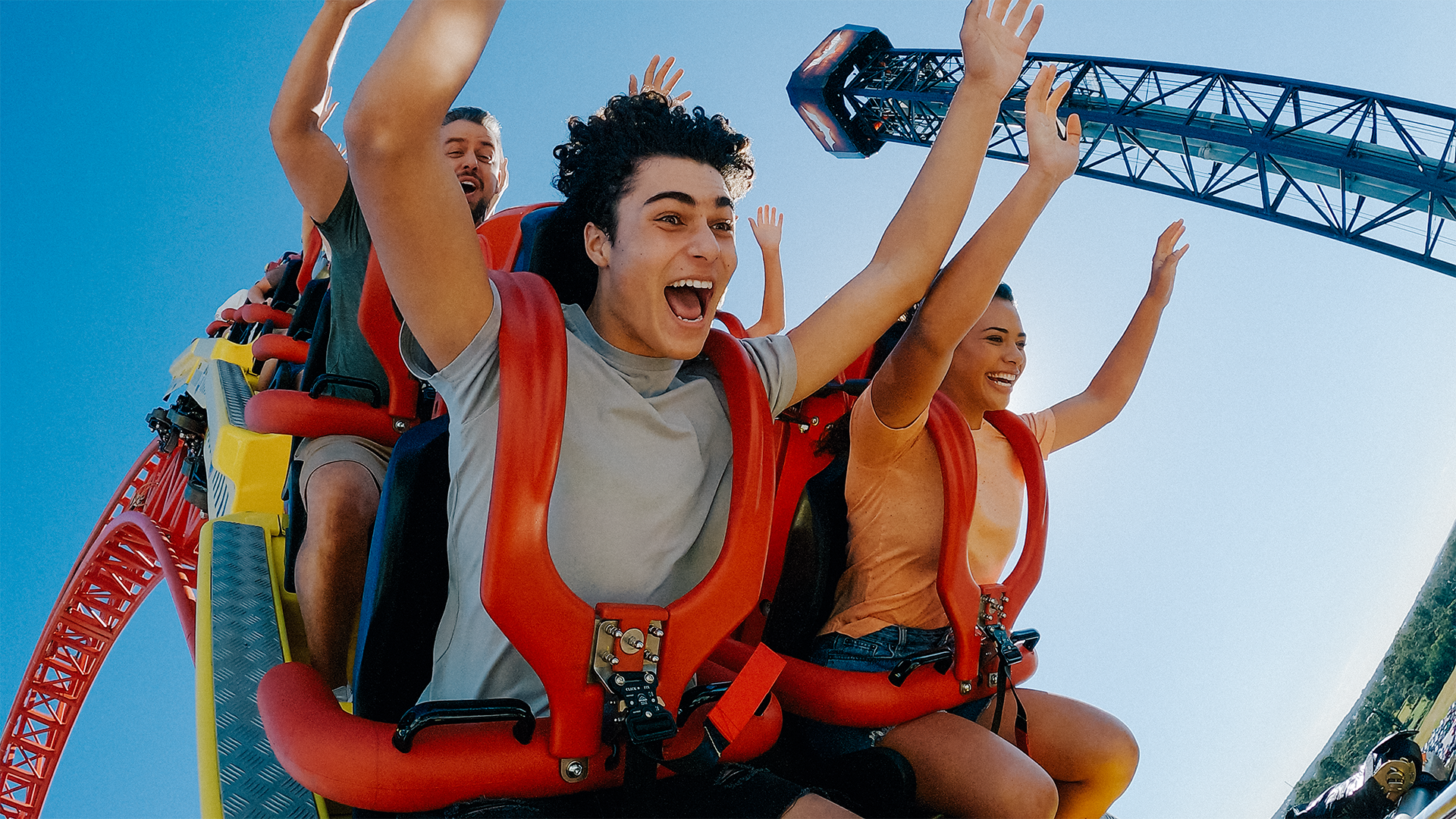 Four people riding a roller coaster with their hands up, smiling and screaming with excitement. The sky is clear and blue, and the roller coaster seats are red.