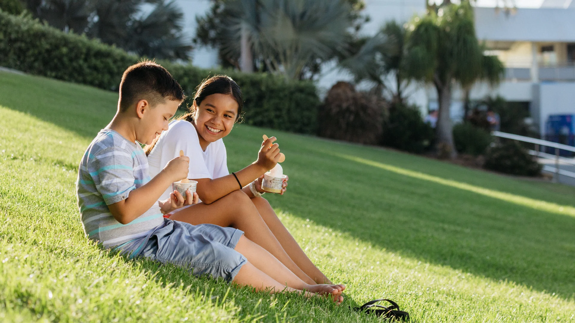 Two children sit on a grassy hill eating ice cream, smiling and facing each other, with trees and a building in the background.