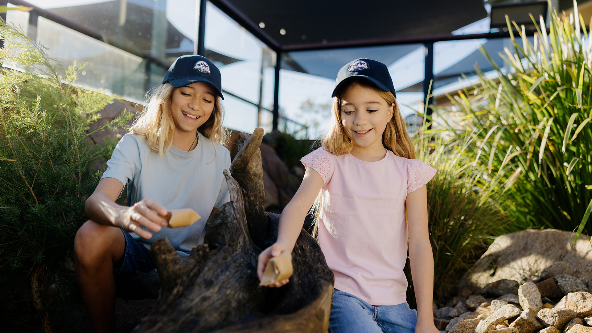 Two young girls wearing navy caps and casual clothes smile while exploring and touching rocks and wood in an outdoor natural setting with greenery and glass walls around them.