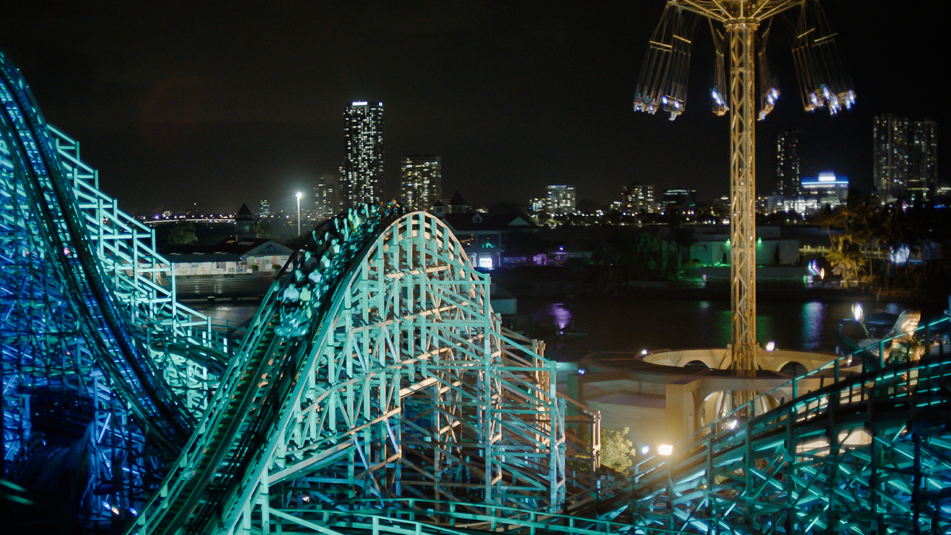 A nighttime cityscape shows a brightly lit roller coaster and a tall swing ride in the foreground, with illuminated buildings and their reflections on water in the background.