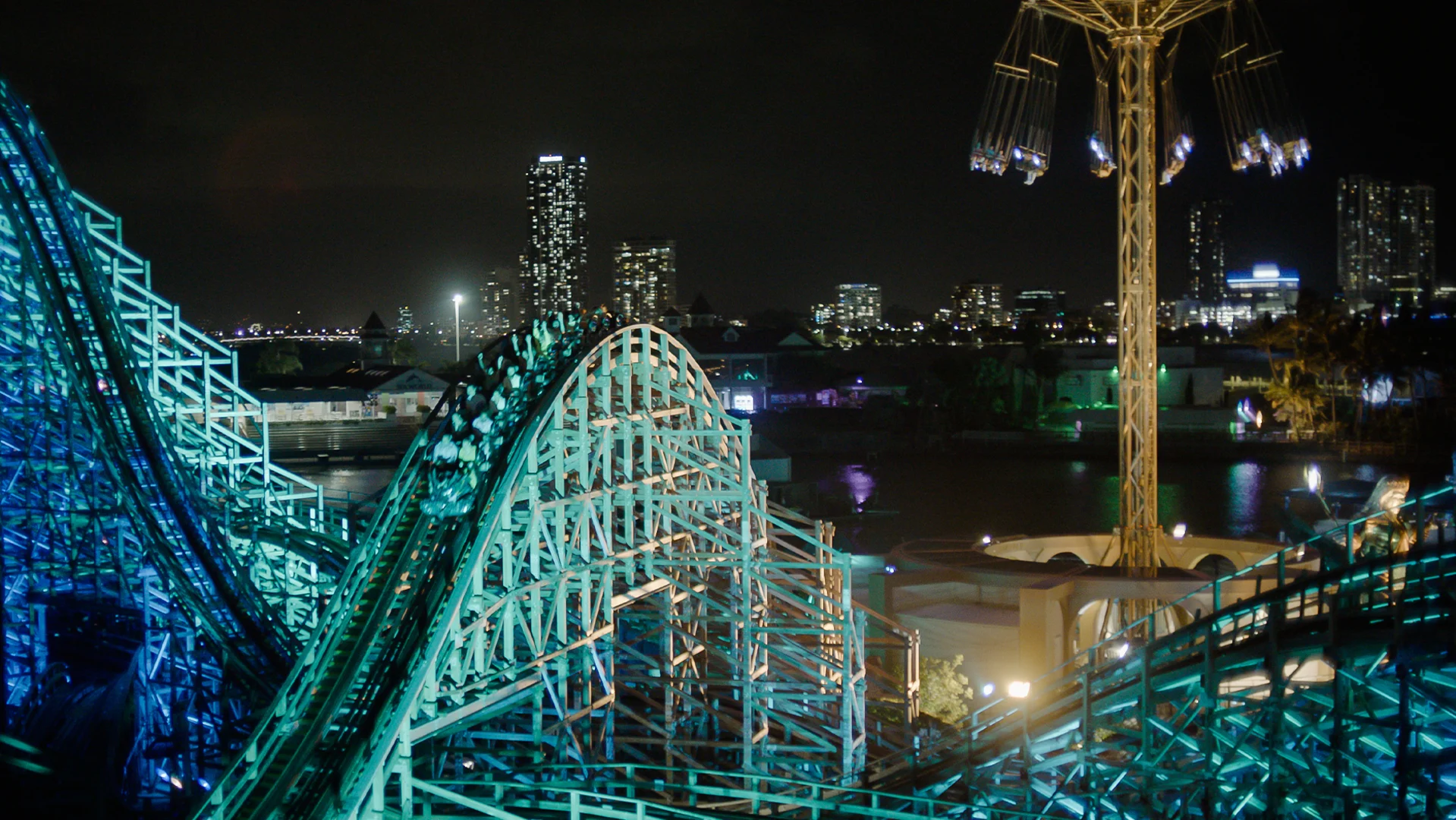 A nighttime cityscape shows a brightly lit roller coaster and a tall swing ride in the foreground, with illuminated buildings and their reflections on water in the background.