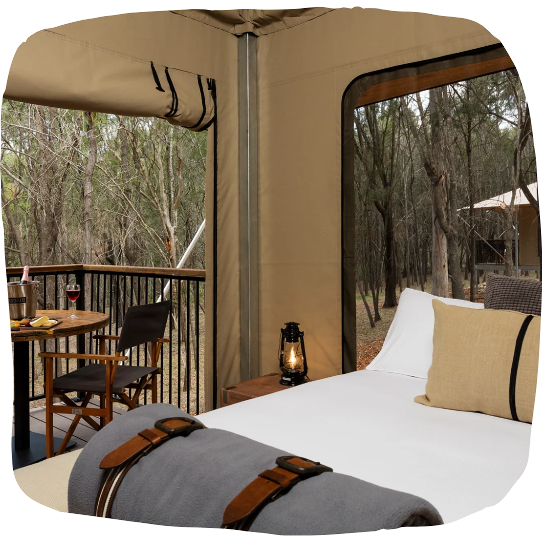 A glamping tent with a neatly made bed, outdoor table and chairs on a deck, lantern, and surrounding forest visible through open sides.