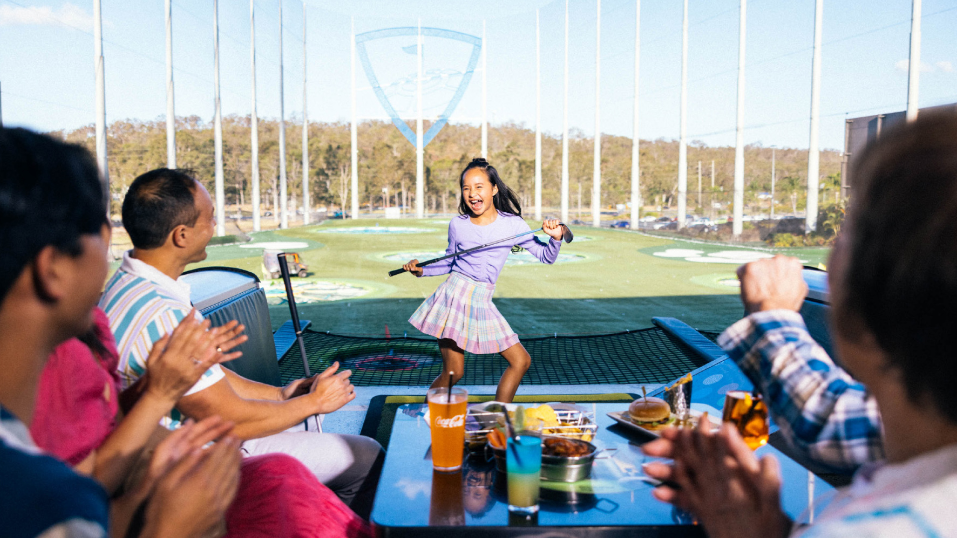 A young girl holding a golf club smiles as a group of people sitting at a table with food and drinks clap for her at a golf driving range.