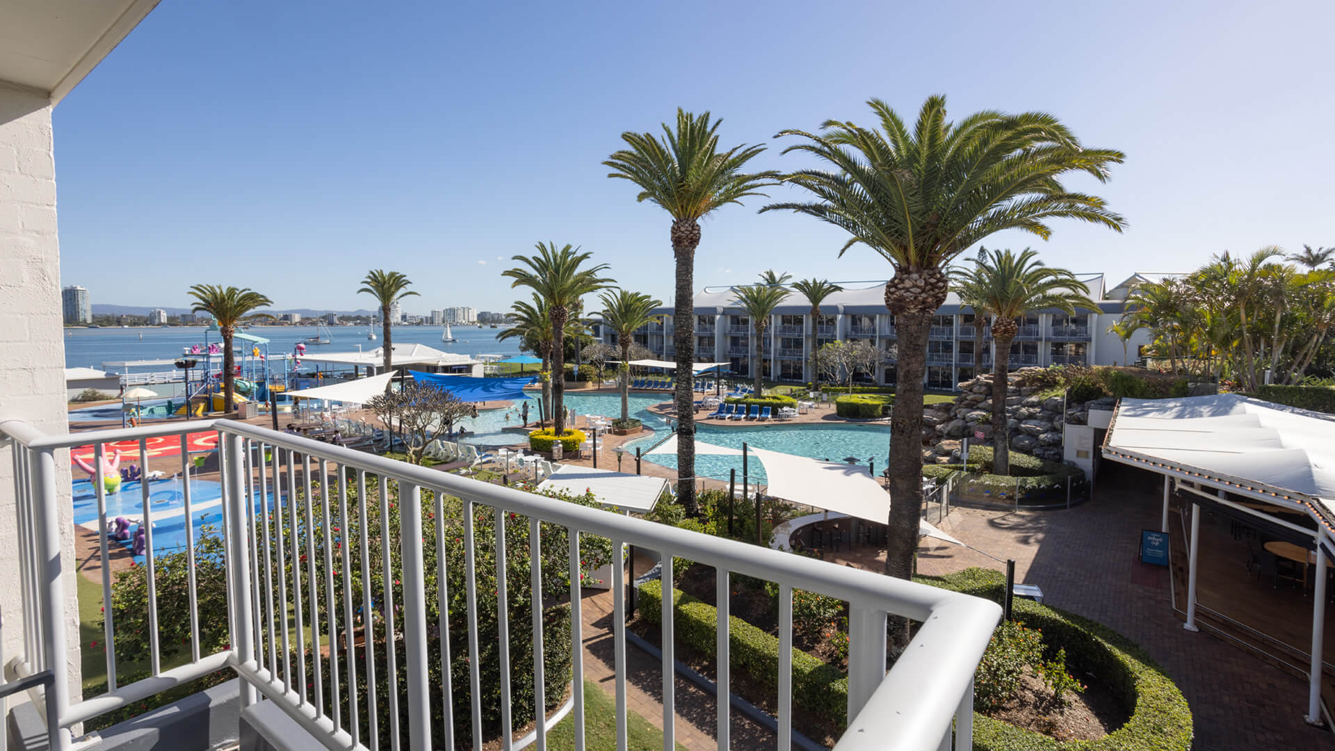 View from a balcony overlooking a hotel pool area with palm trees, lounge chairs, and buildings in the background under a clear blue sky.