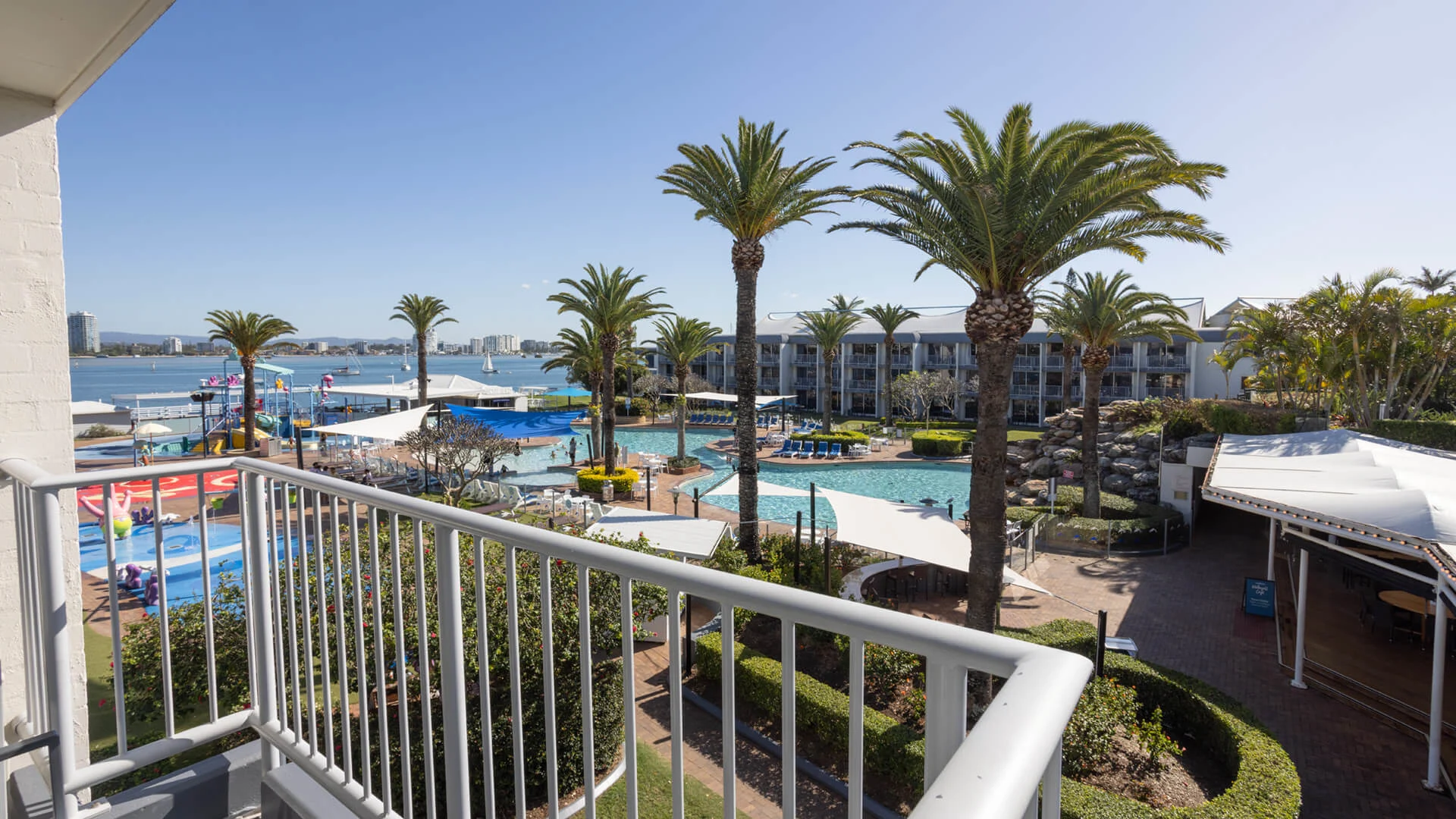 View from a balcony overlooking a hotel pool area with palm trees, lounge chairs, and buildings in the background under a clear blue sky.