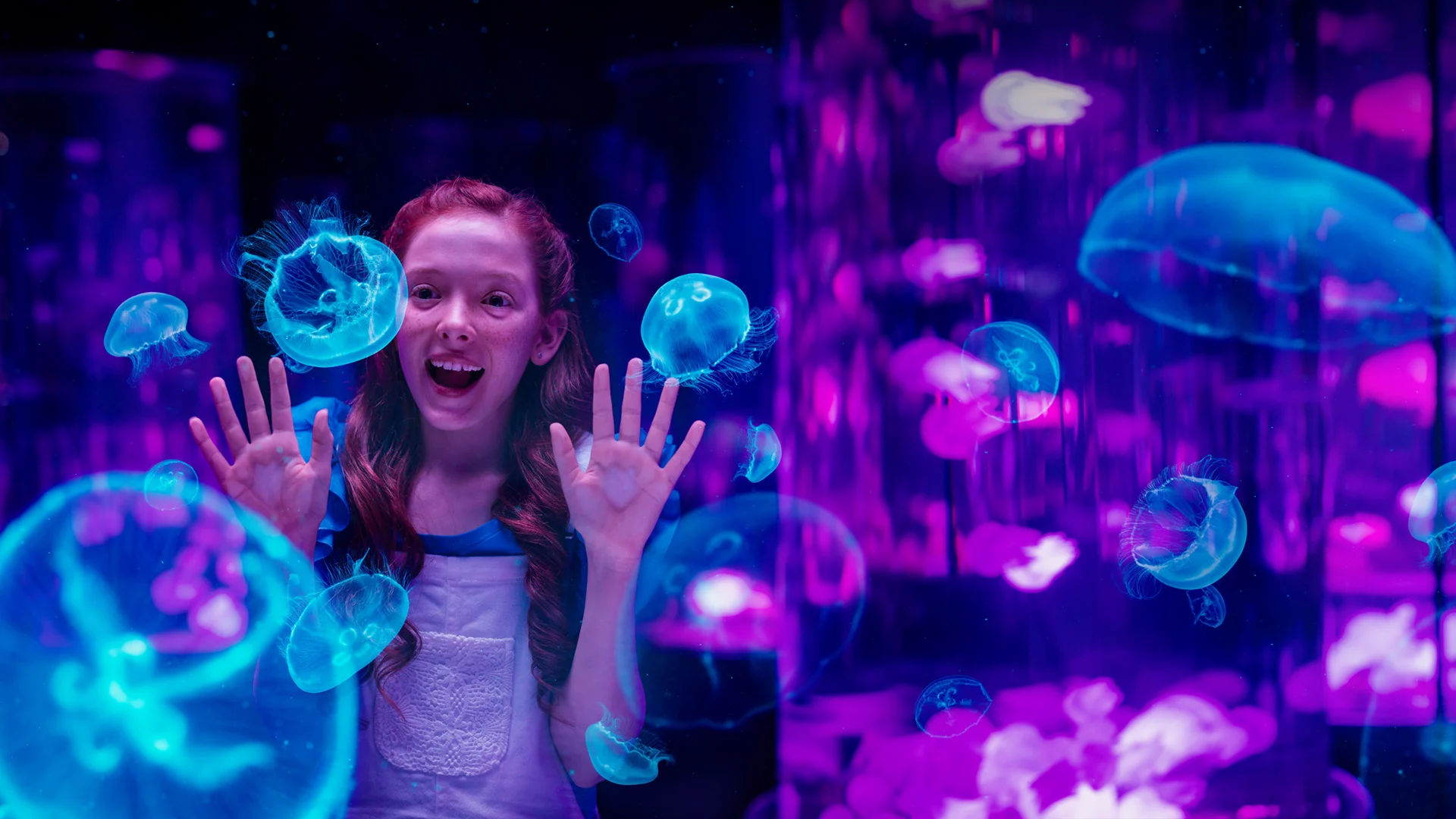 A girl with long red hair looks excited as she observes blue jellyfish behind glass in a dimly lit, purple-accented aquarium.