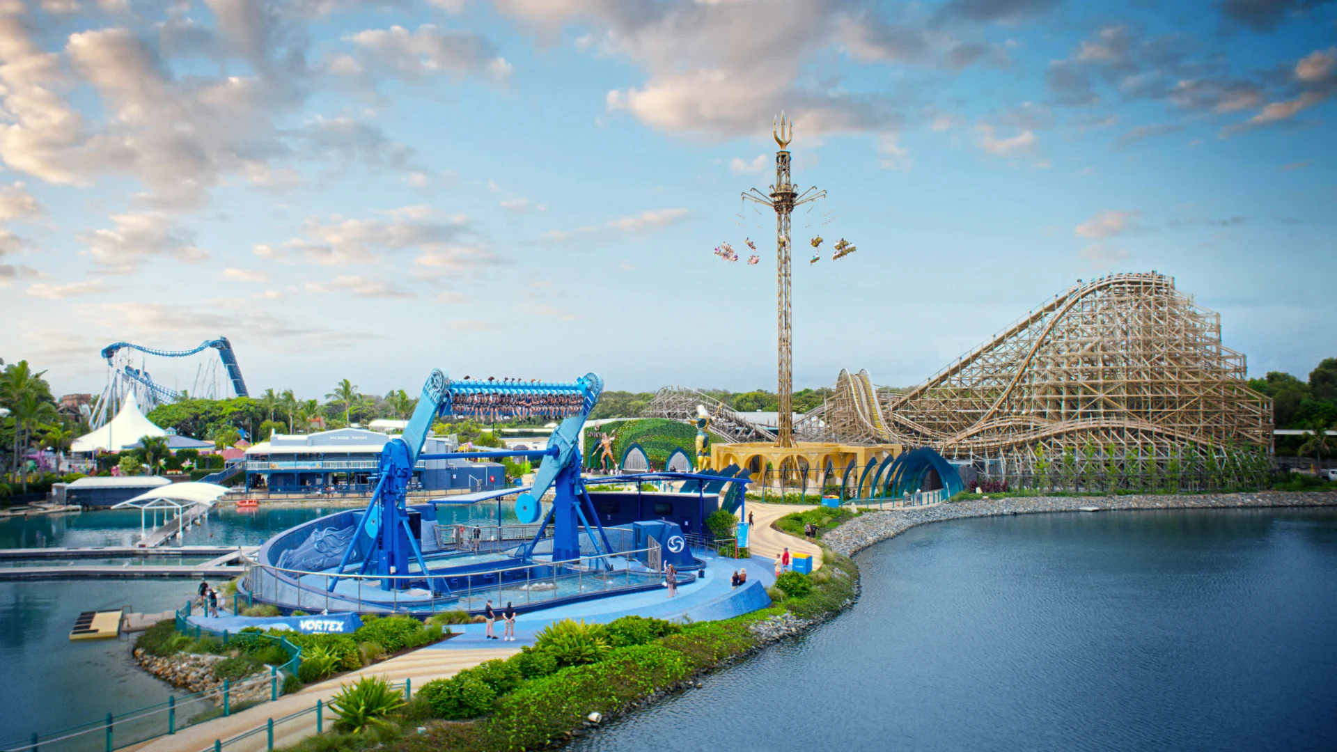 A theme park scene featuring a wooden roller coaster, a blue stadium, a tall spinning ride, and pathways around a body of water under a partly cloudy sky.