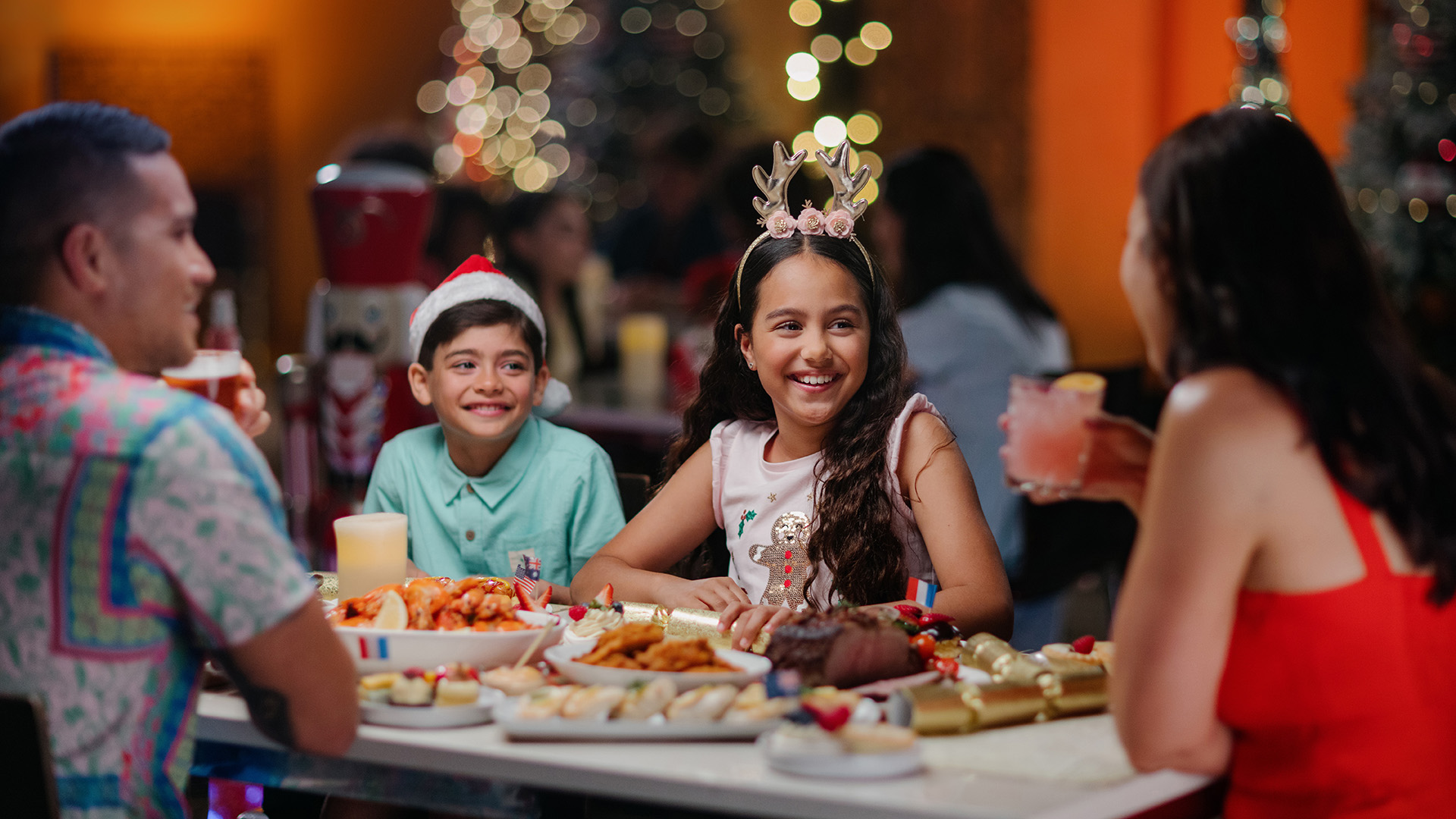 A family of four sits at a festive dinner table, decorated with holiday treats. Two children wear holiday headbands and smile, while parents talk and enjoy drinks. Twinkling Christmas lights and a tree glow in the background.