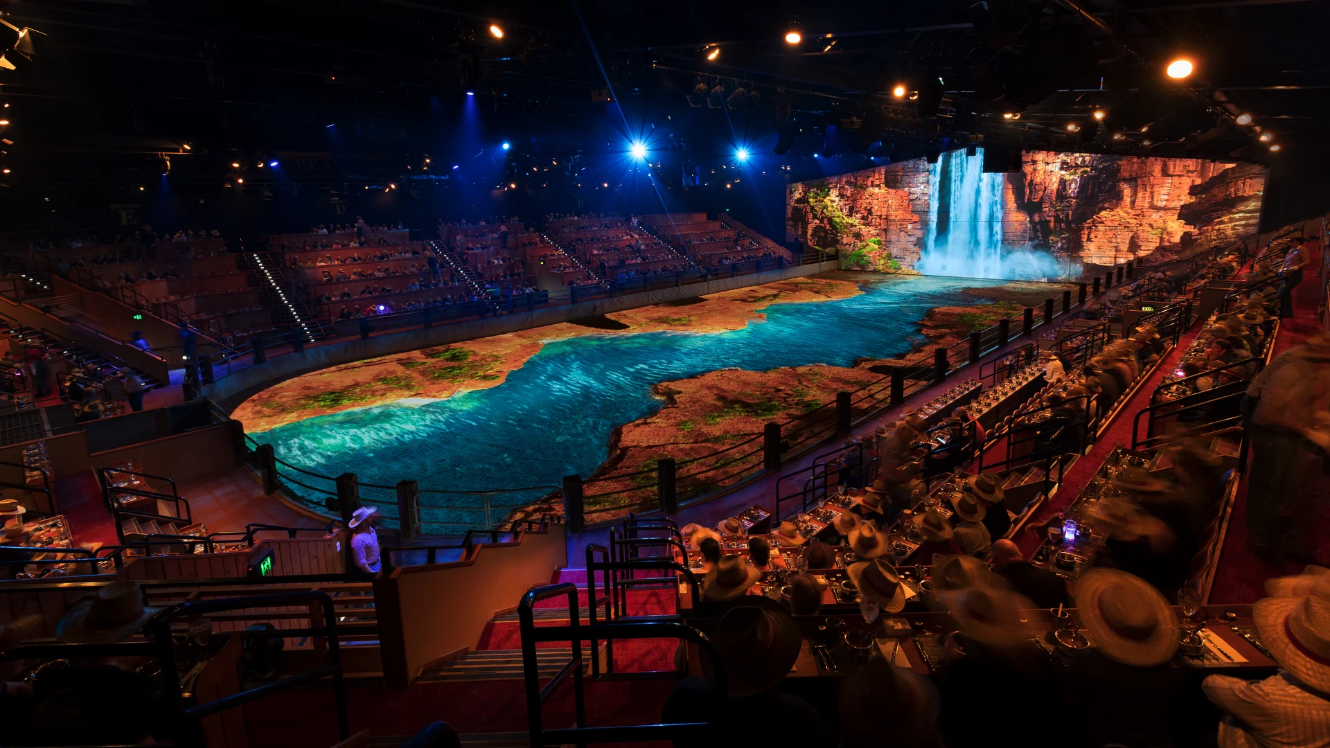 A large indoor arena with audience seating surrounds a stage designed to look like a river with flowing water and a scenic waterfall projected on a screen in the background, under dramatic stage lighting.