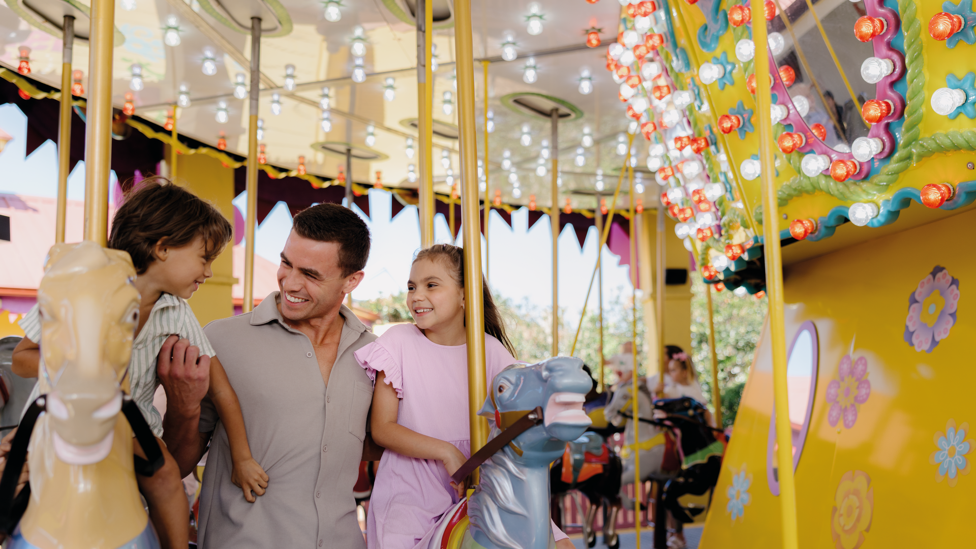 A man with a young boy and girl smile together while standing next to colorful carousel horses on a brightly lit, decorated carousel ride at an amusement park.
