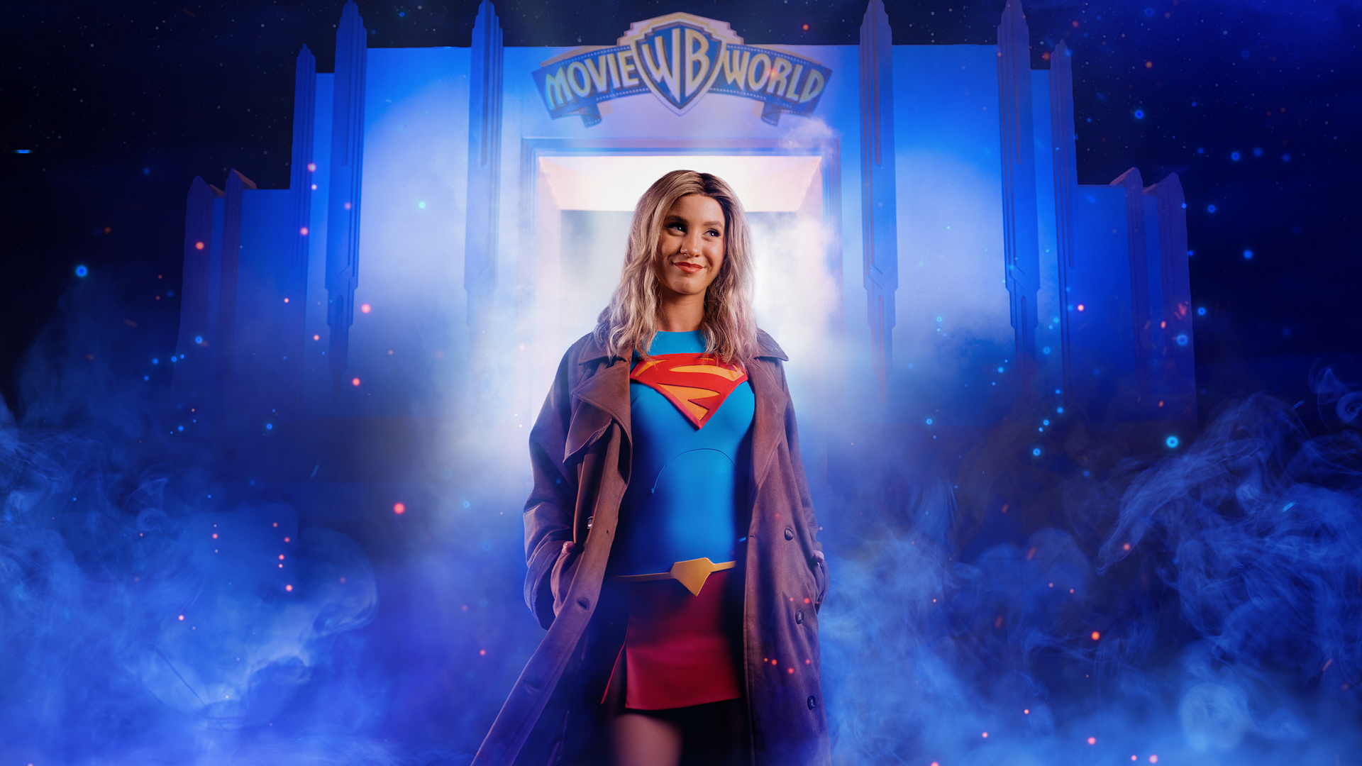 A woman dressed as Supergirl stands confidently in front of the glowing entrance to "WB Movie World," surrounded by blue mist and dramatic lighting.