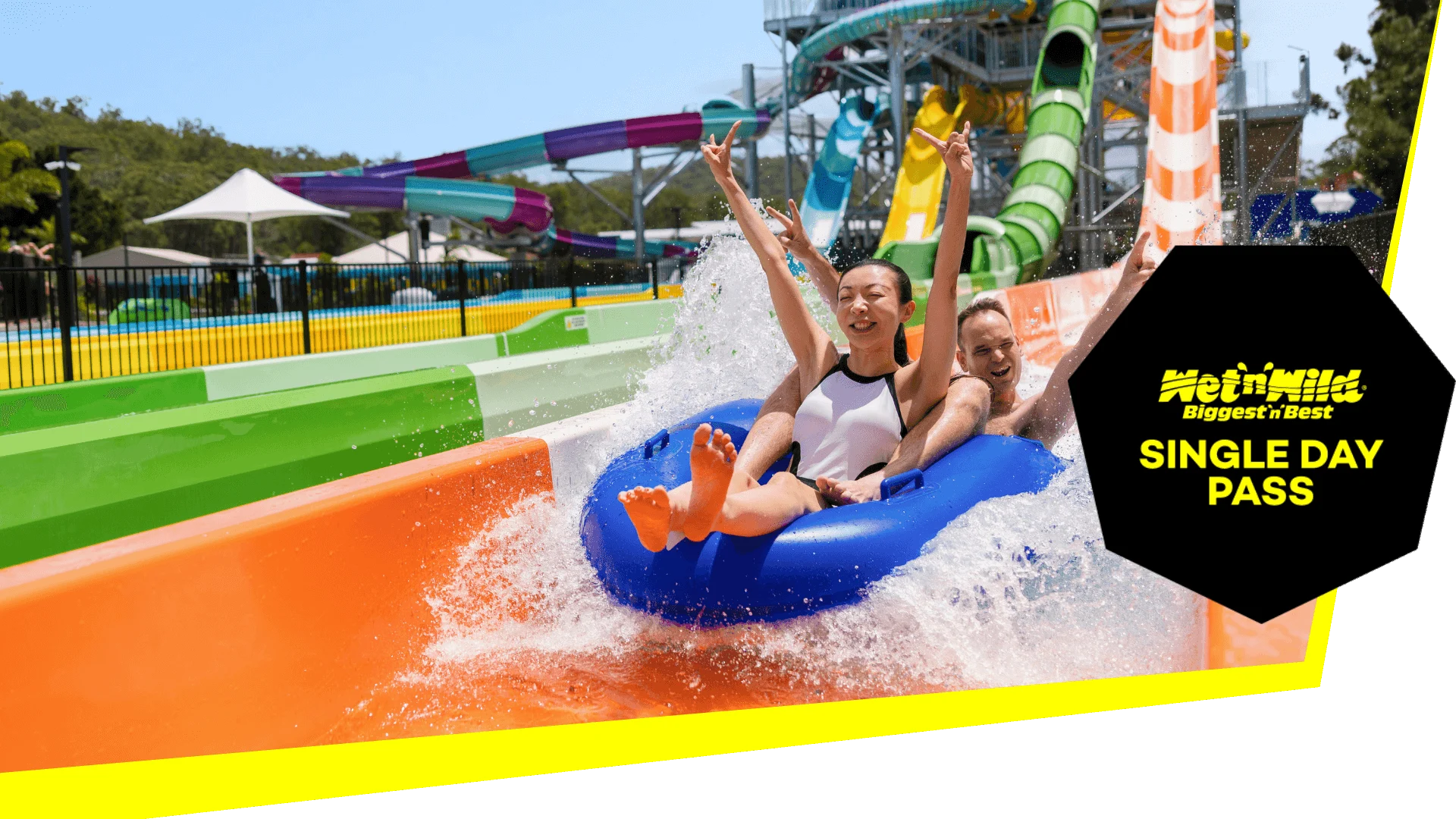 Two people on a blue inflatable raft enjoy a thrilling water slide ride at the water park. The image text reads, "Wet'n'Wild Biggest 'n' Best, Single Day Ticket.