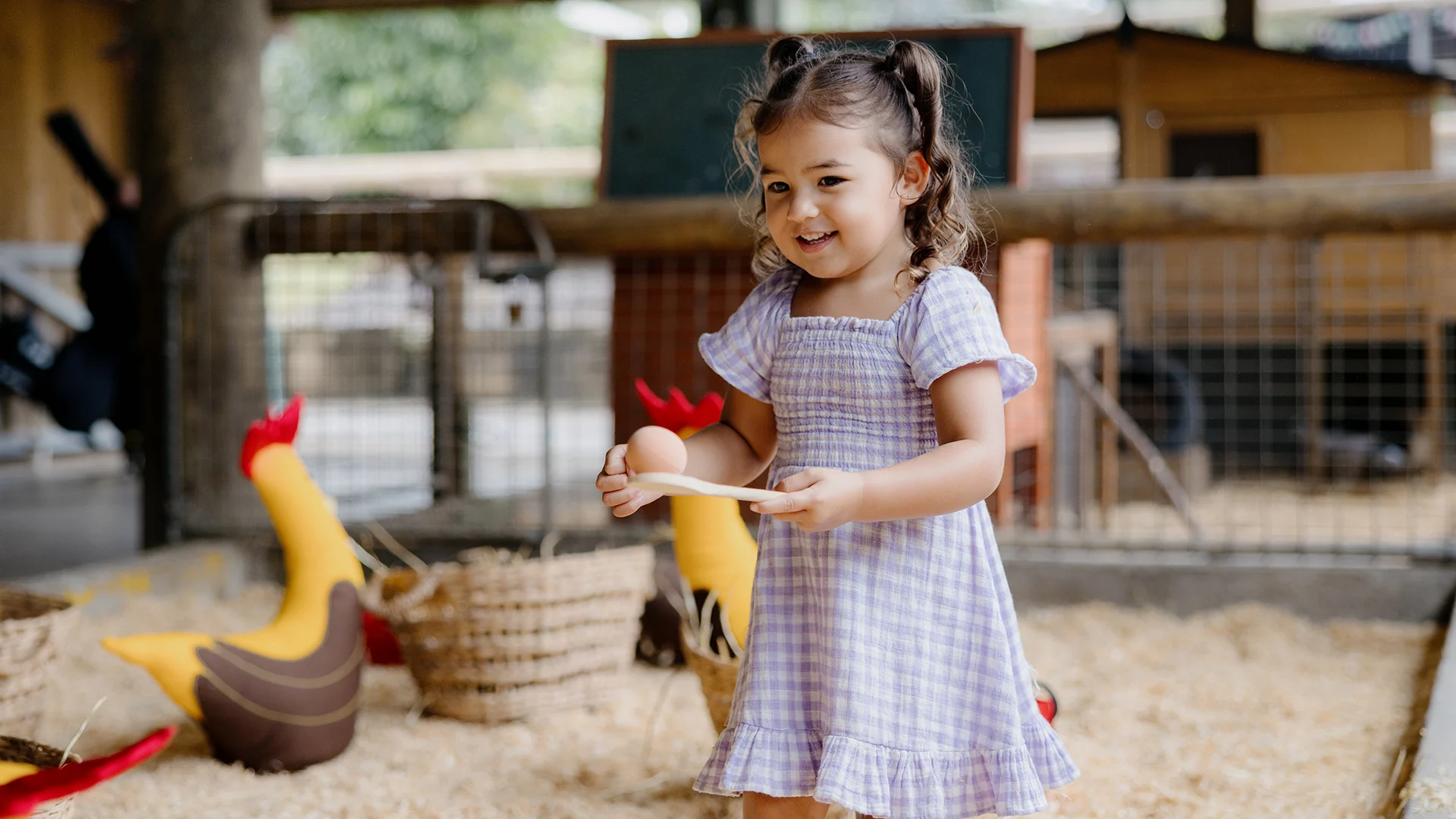 A young girl in a purple checkered dress smiles while holding a wooden egg on a spoon, standing in a barn play area with toy chickens and straw.