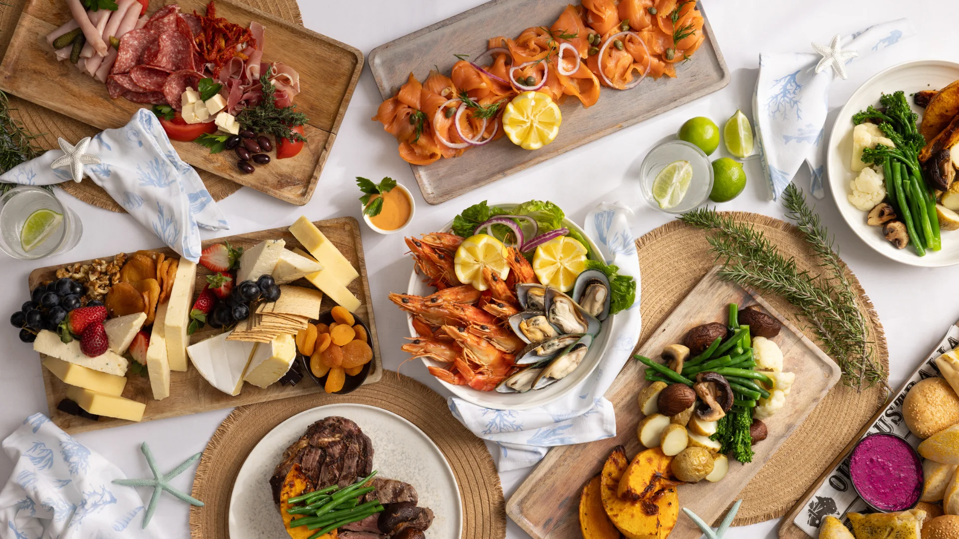 An overhead view of a variety of dishes on a table, including seafood, meats, cheeses, fruits, and breads, arranged on wooden boards and plates with decorative elements and napkins.