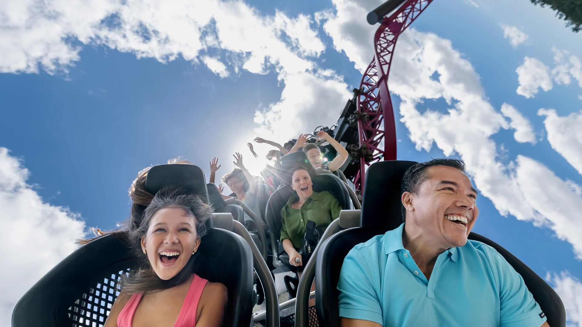 A group of people smiling and laughing as they ride a roller coaster under a bright blue sky with scattered clouds. The track rises steeply behind them, capturing their excitement and thrill.