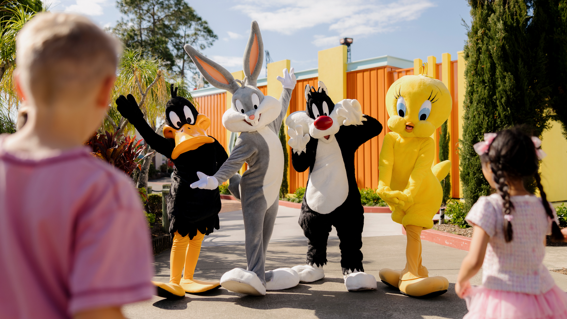 Four children approach people in costumes of Daffy Duck, Bugs Bunny, Sylvester, and Tweety Bird outside on a sunny day, with colorful buildings and trees in the background.
