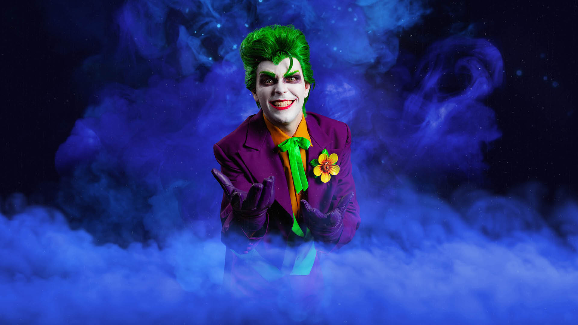 A person dressed as the Joker, with green hair, white face makeup, and a purple suit with a yellow shirt and green tie, stands in front of swirling blue smoke, smiling mischievously.
