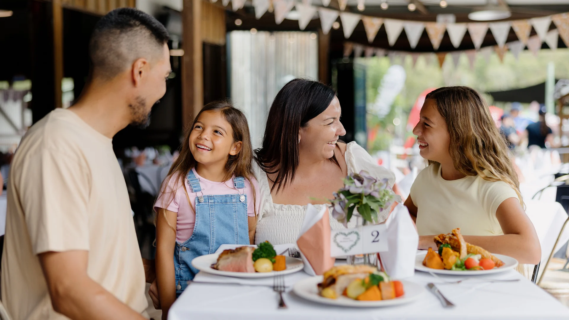 A family of four—two adults and two young girls—sit at a restaurant table outdoors, smiling and enjoying a meal together. The table is set with plates of food and a small flower arrangement.