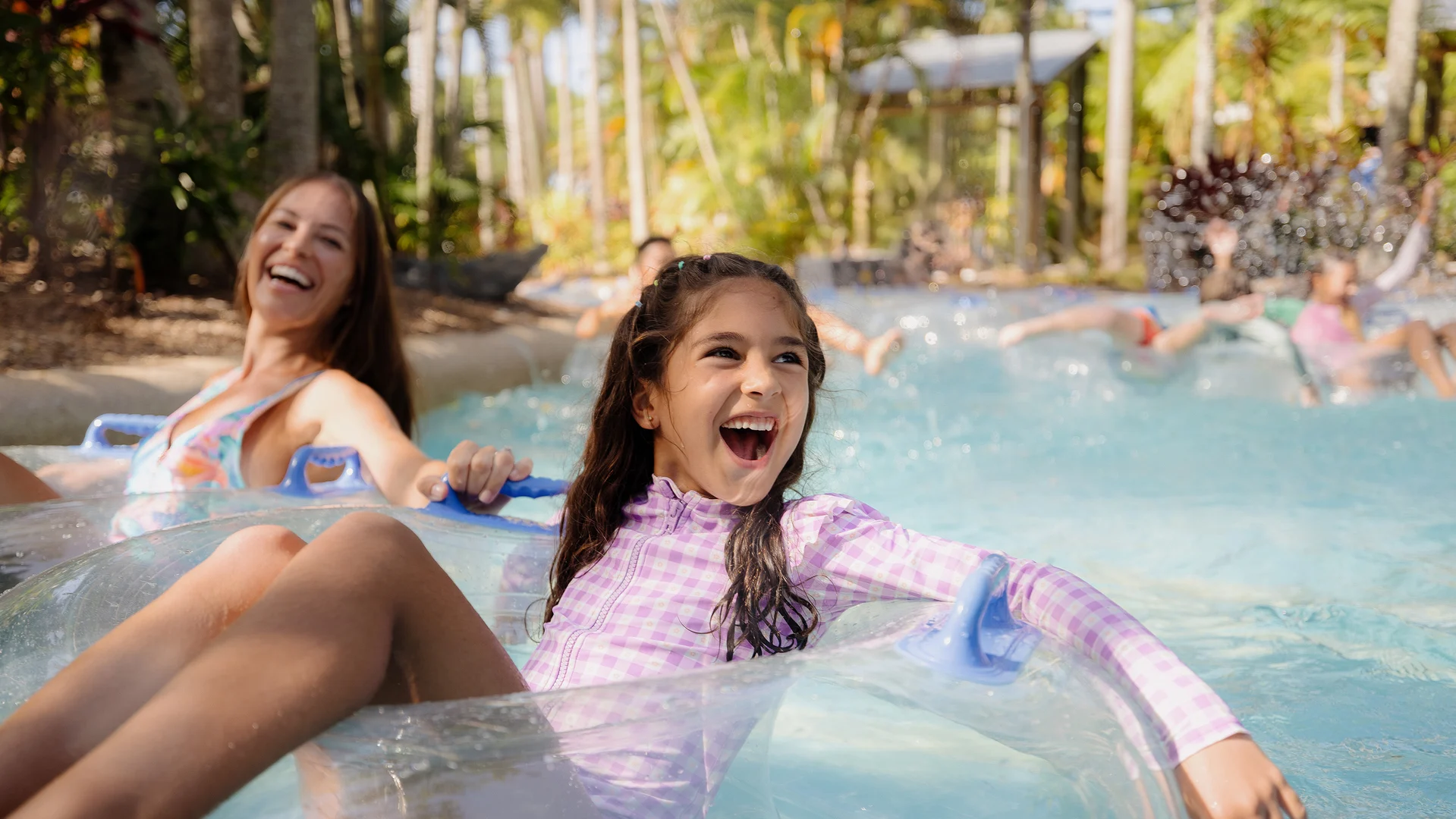Two girls are laughing and relaxing on clear inflatable tubes in a lazy river at a water park, surrounded by palm trees and greenery under bright sunlight. Other people float in the background.