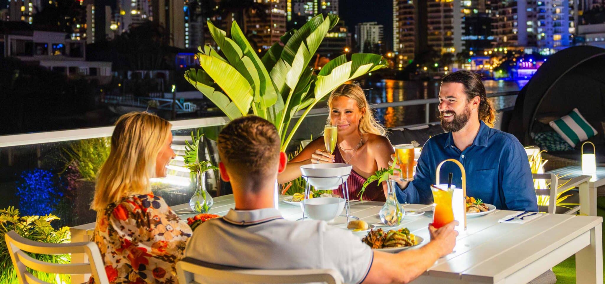 Four people sit at an outdoor restaurant table at night, enjoying food and drinks, with city buildings and lights in the background.