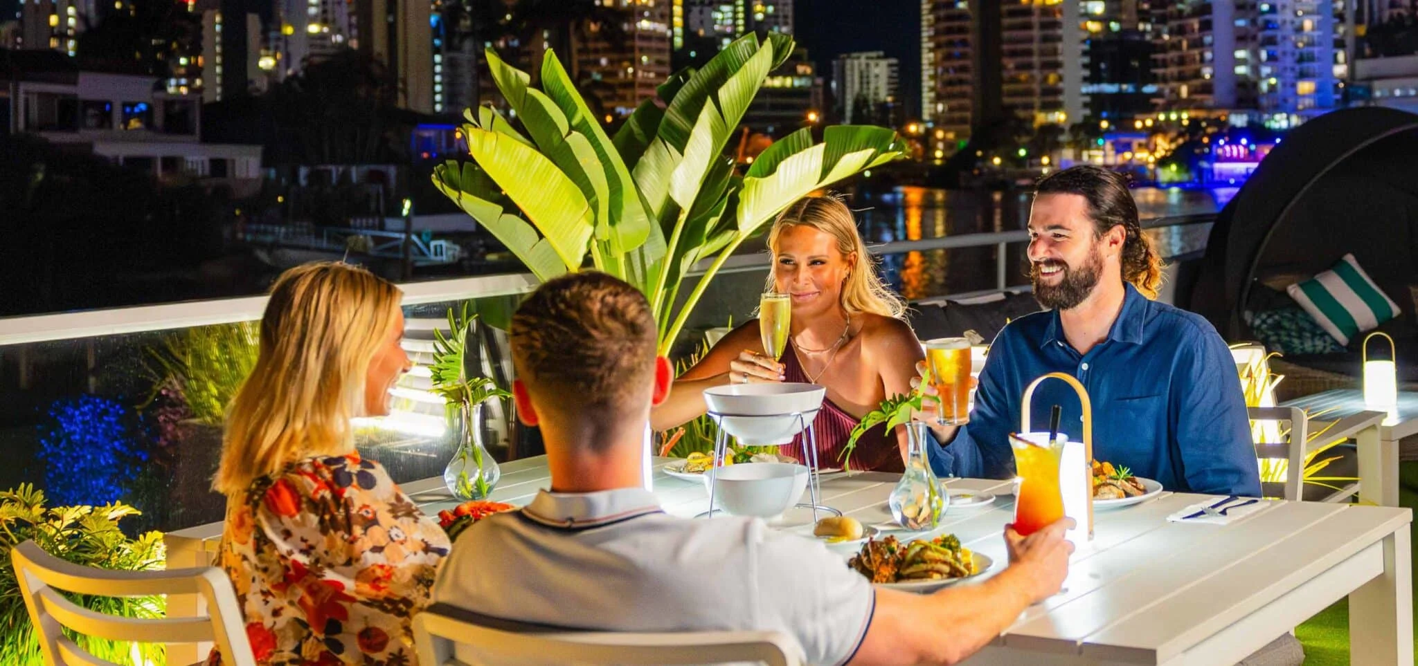Four people sit at an outdoor restaurant table at night, enjoying food and drinks, with city buildings and lights in the background.