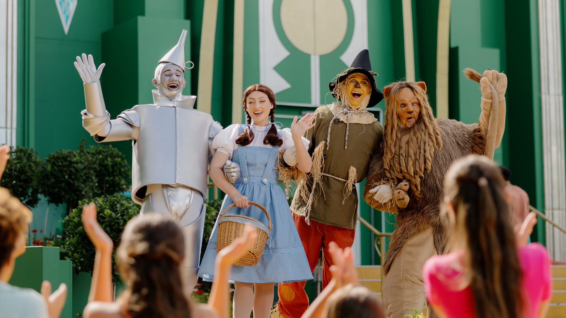 Four people dressed as the Tin Man, Dorothy, Scarecrow, and Cowardly Lion from “The Wizard of Oz” stand smiling and waving on stage, as children in the audience clap and cheer.