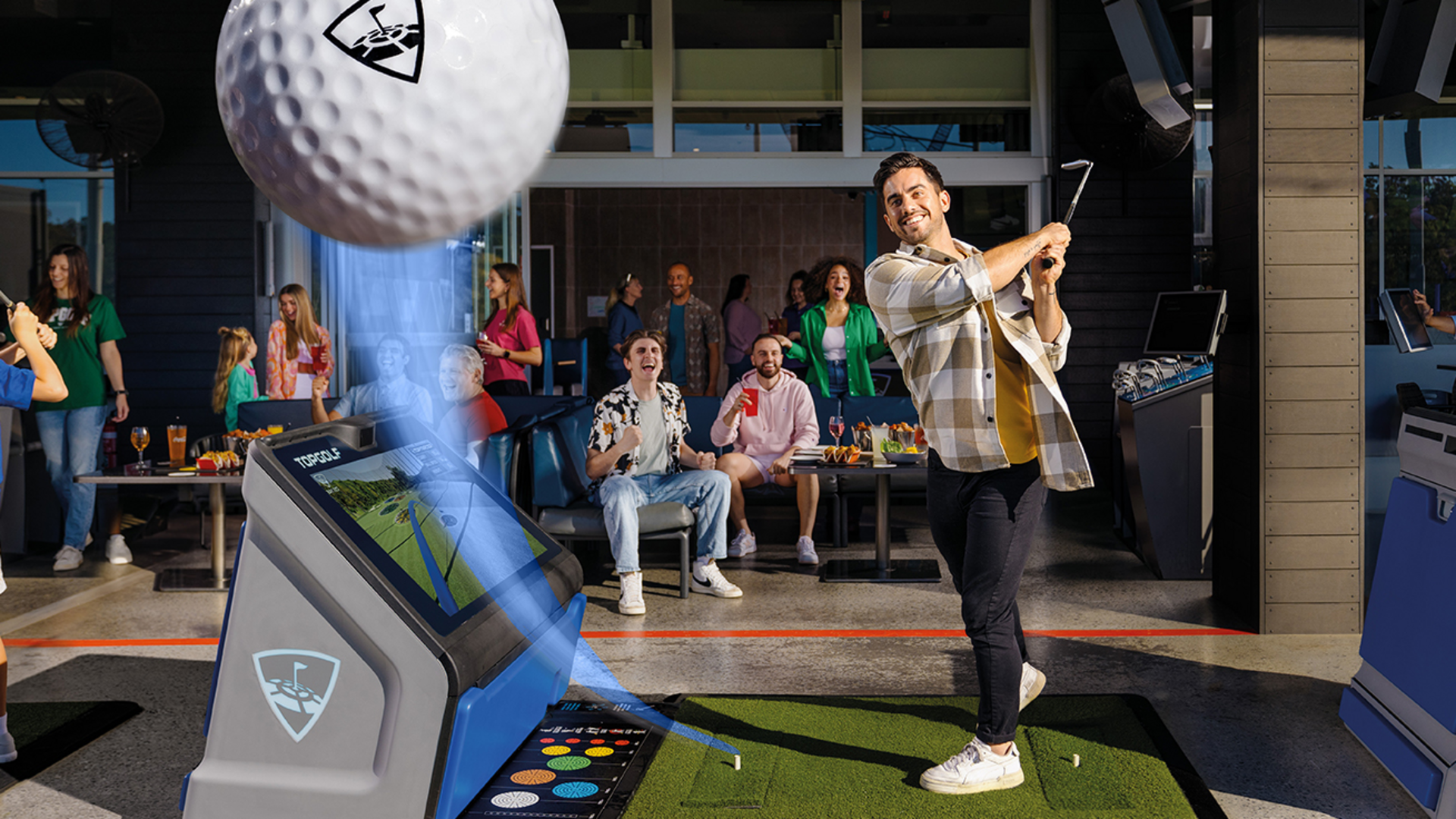 A man swings a golf club at an indoor driving range while a large golf ball appears in the foreground. Several people sit and cheer in the background, enjoying drinks and watching the action.