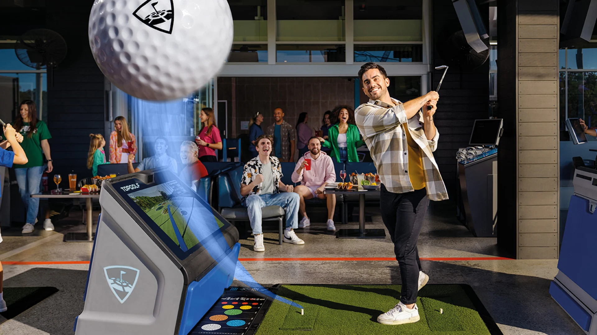 A man swings a golf club at an indoor driving range while a large golf ball appears in the foreground. Several people sit and cheer in the background, enjoying drinks and watching the action.