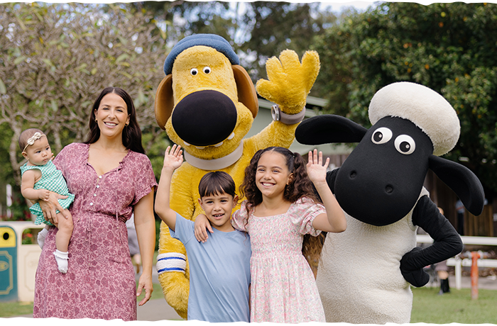 A woman holding a baby stands beside two children, all smiling and waving, with large dog and sheep mascots waving behind them in an outdoor park setting.