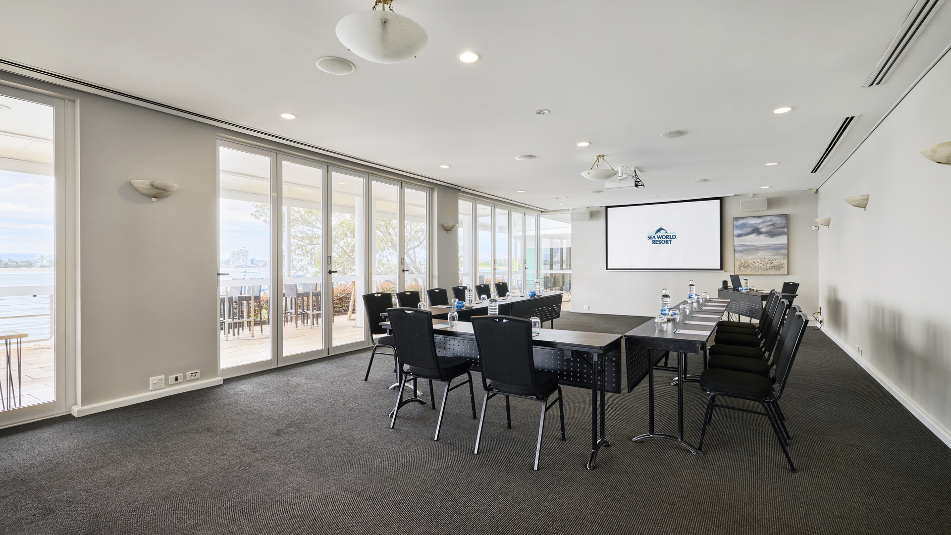 A modern conference room with black chairs arranged around square tables, a large projector screen at the front, glass doors leading to a balcony with a view, and bright natural light filling the space.