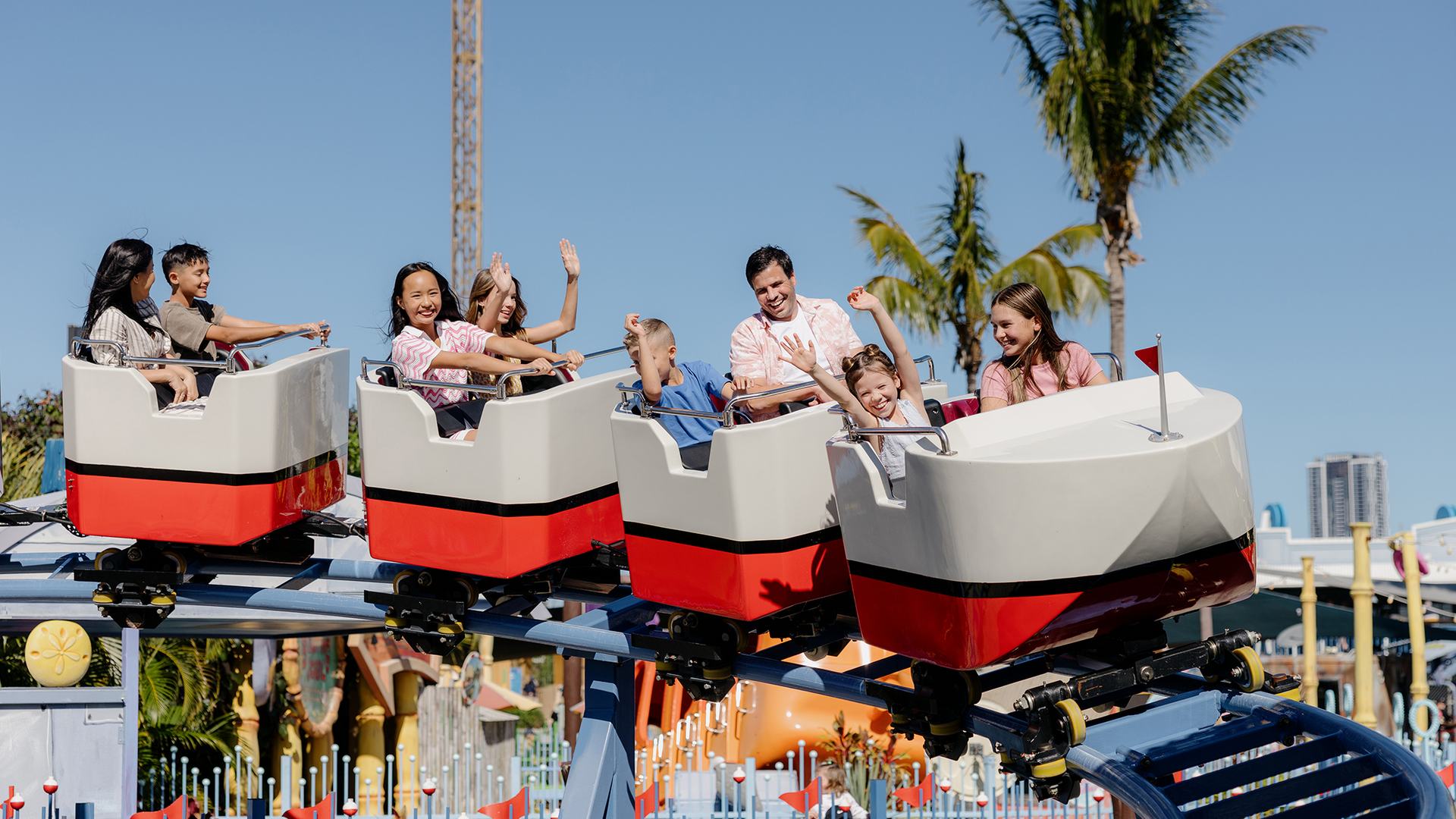 Children and adults ride a small roller coaster with white and red cars at an amusement park on a sunny day. Palm trees and city buildings are visible in the background.