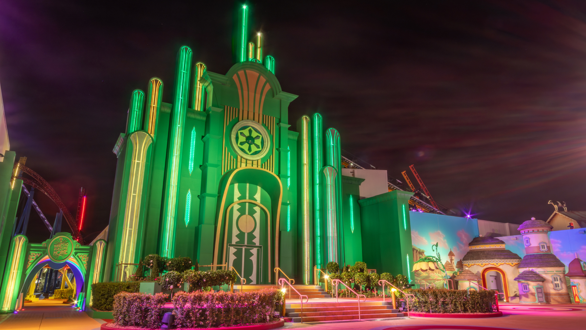A building resembling the Emerald City from The Wizard of Oz, glowing with bright green and yellow neon lights at night, with steps leading to a large arched door and whimsical structures nearby.