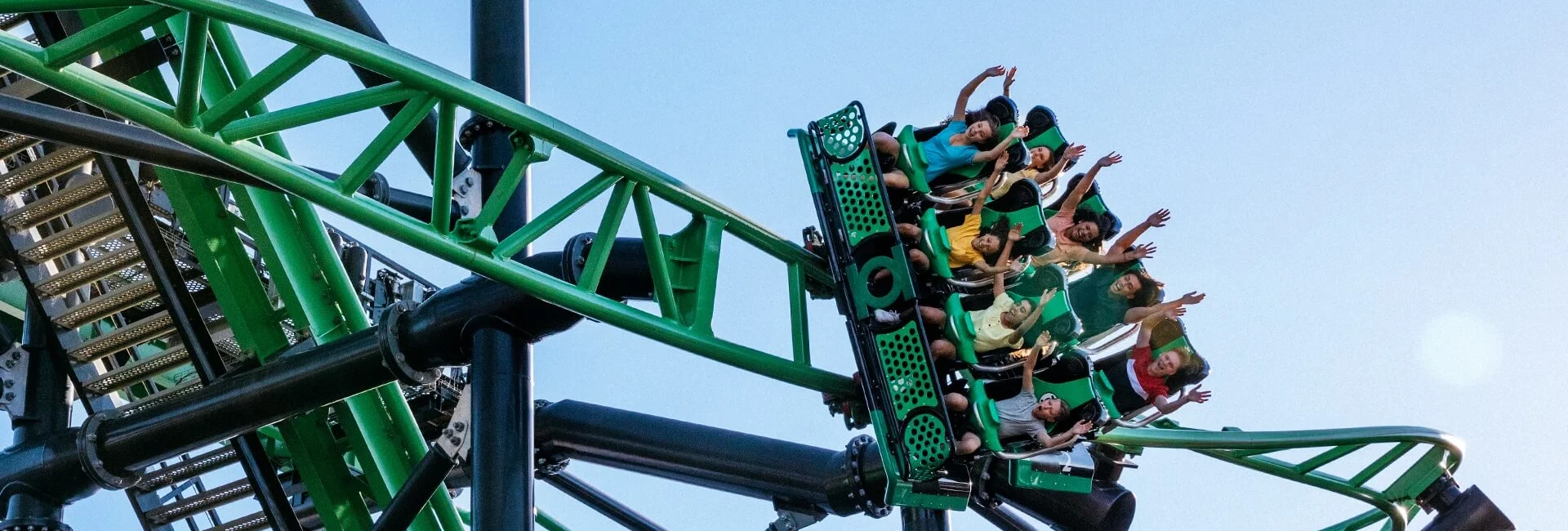 A group of people ride a green and black roller coaster with their arms raised, against a clear blue sky background.