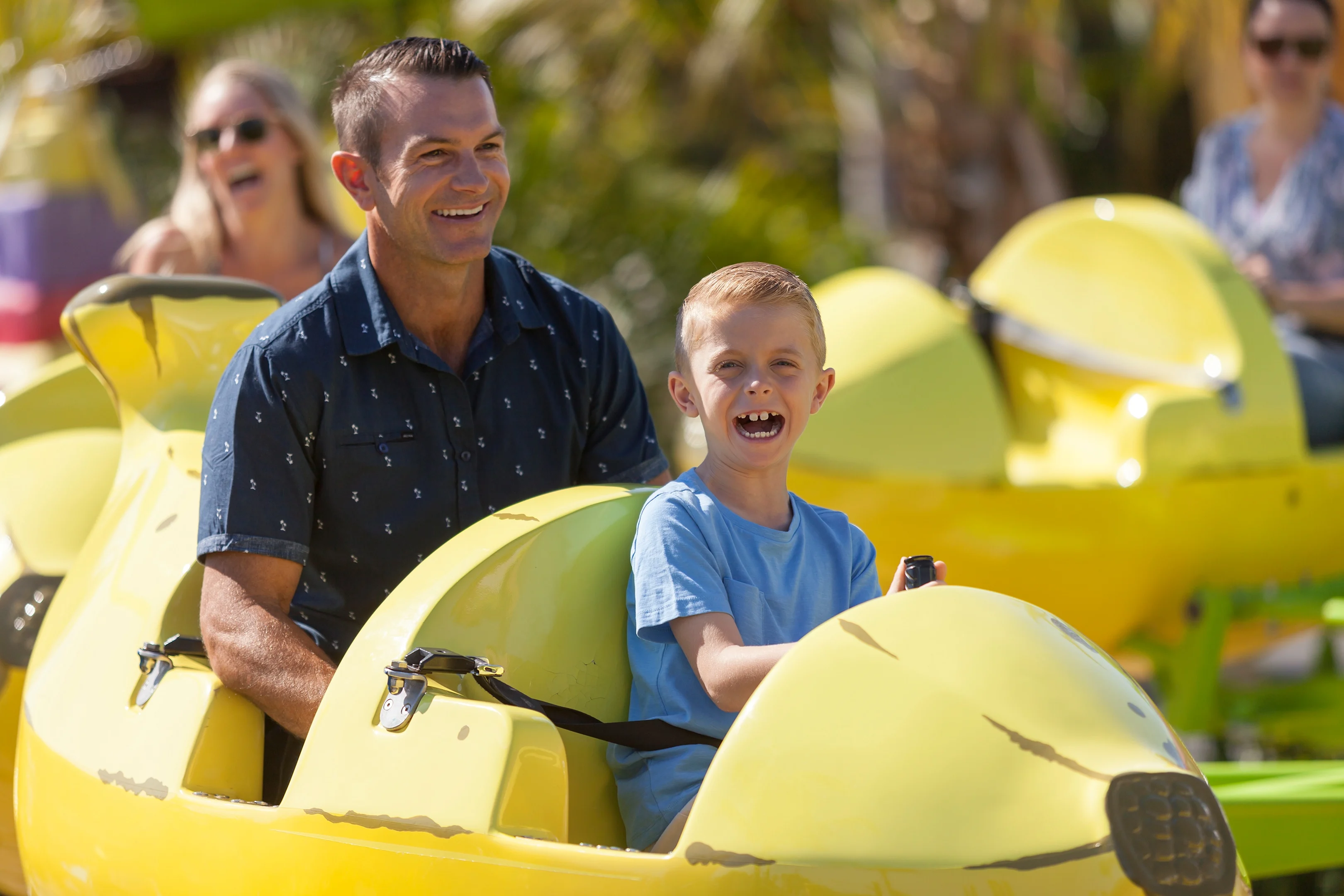 A man and a boy ride together in a yellow amusement park ride shaped like an airplane, both smiling. Other people are visible in the background.