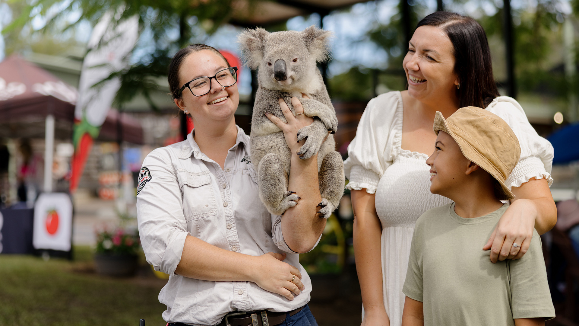 Two women and a child smile outdoors as one woman holds a koala. A market with tents and banners is visible in the background, and the group appears happy and relaxed.