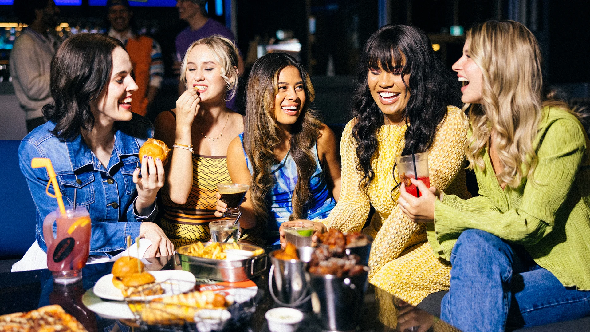 Five women sit together at a table, laughing and enjoying drinks and food. The table is filled with burgers, fries, and cocktails. The atmosphere is lively and friendly, suggesting a fun social gathering.