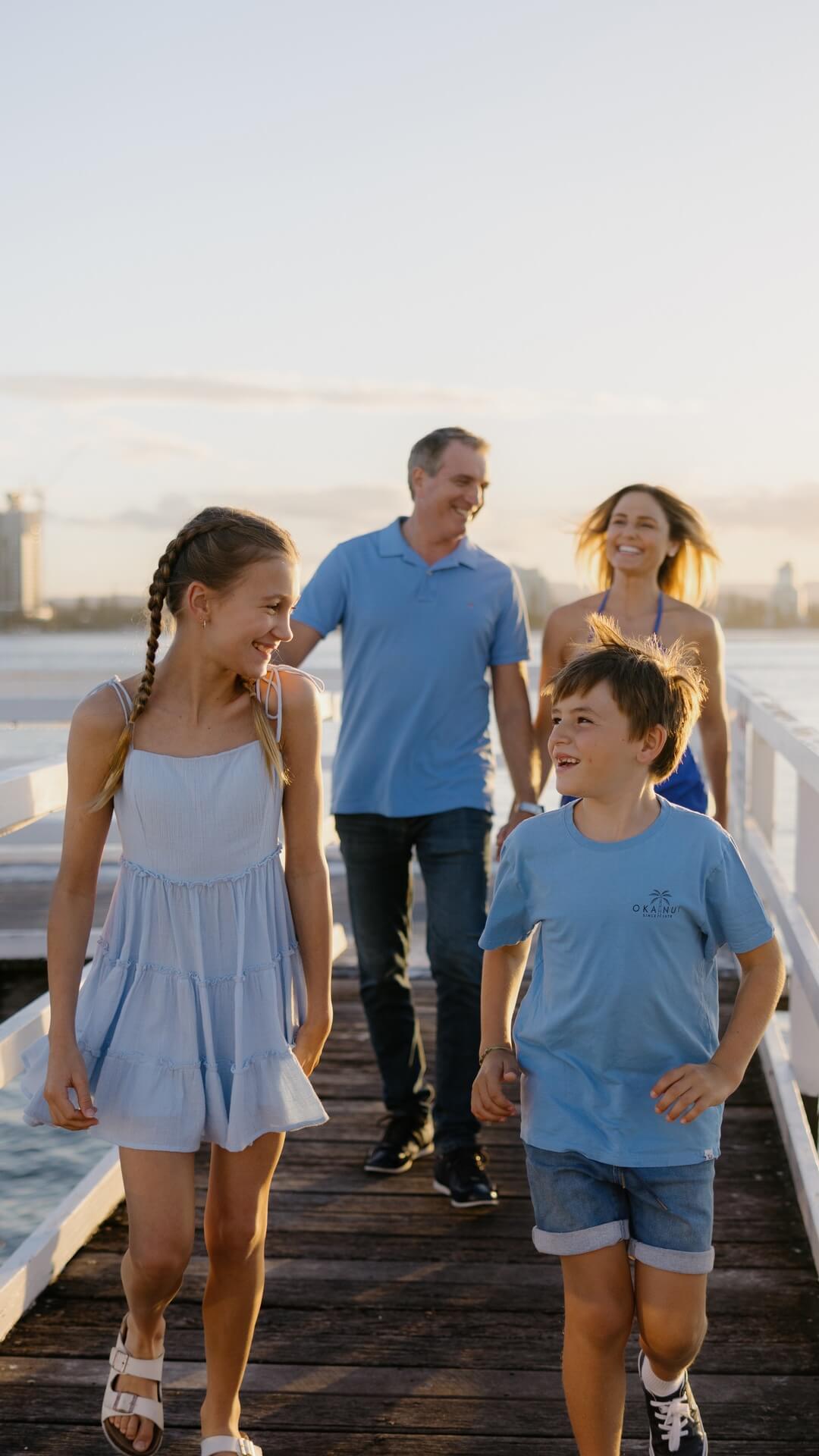 Family of four walking on a wooden pier at sunset. Two children in the front, flanked by two adults, all smiling and wearing casual summer outfits. Water and city skyline in the background.