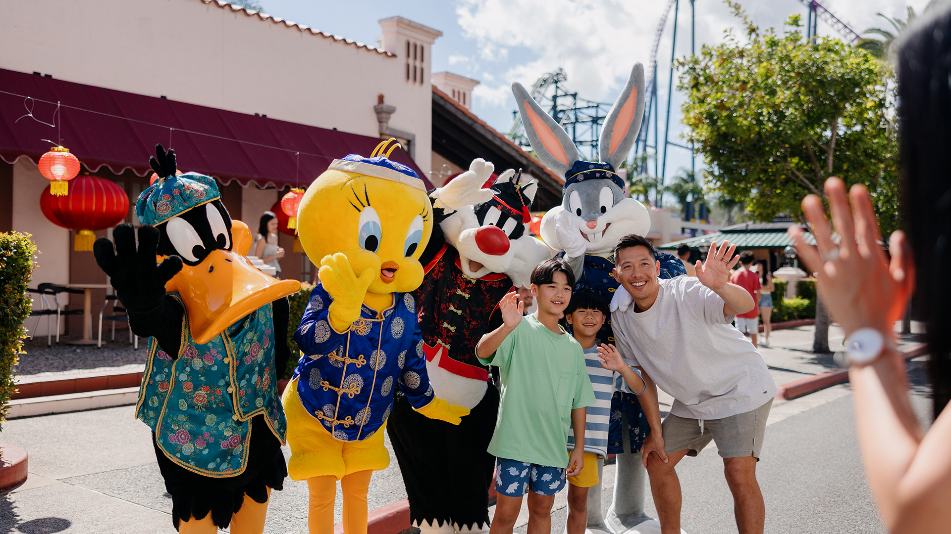A group of children and an adult pose and wave for a photo with people dressed as Looney Tunes characters—Daffy Duck, Tweety, Sylvester, and Bugs Bunny—at an outdoor amusement park.
