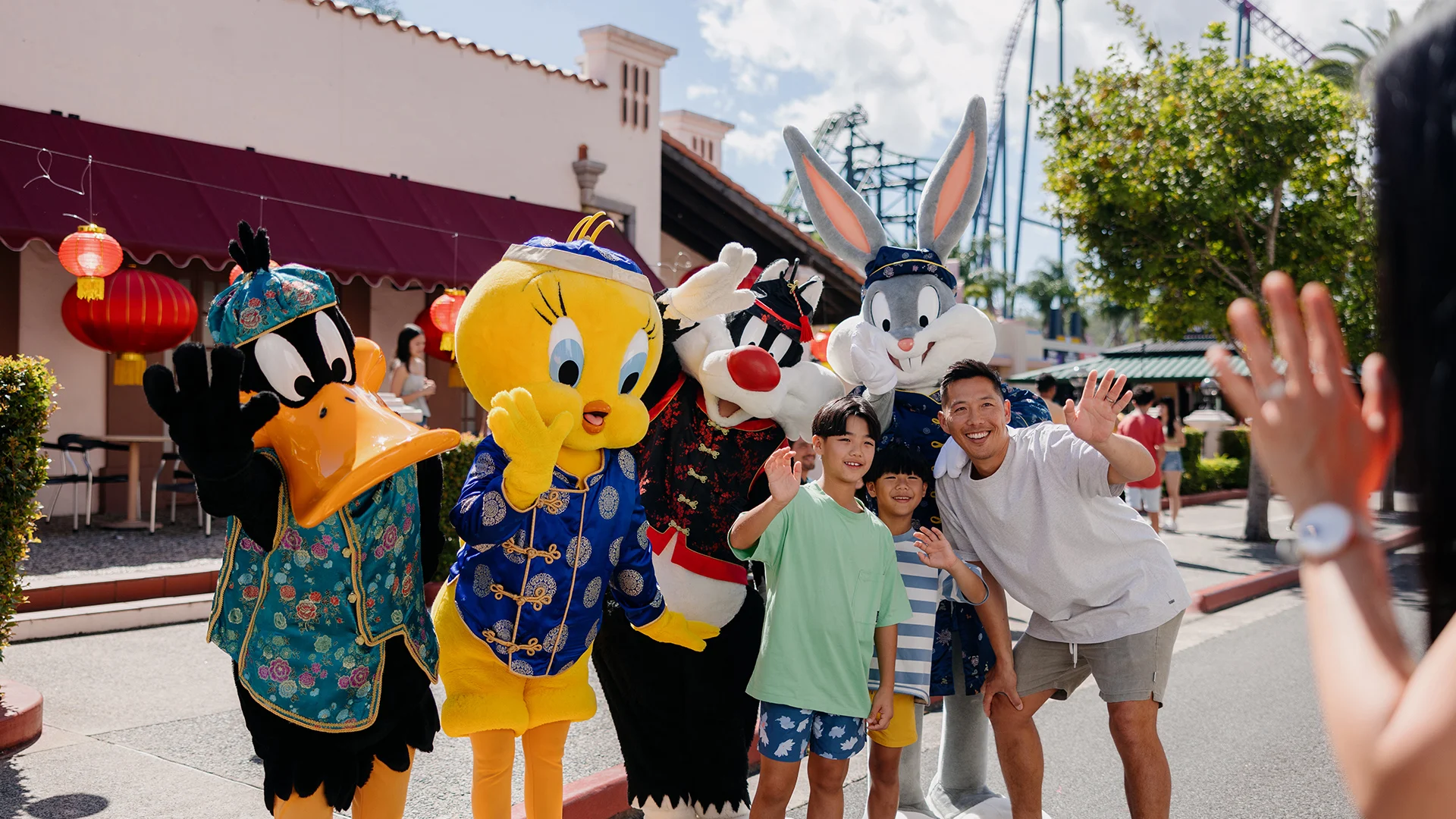 A group of children and an adult pose and wave for a photo with people dressed as Looney Tunes characters—Daffy Duck, Tweety, Sylvester, and Bugs Bunny—at an outdoor amusement park.