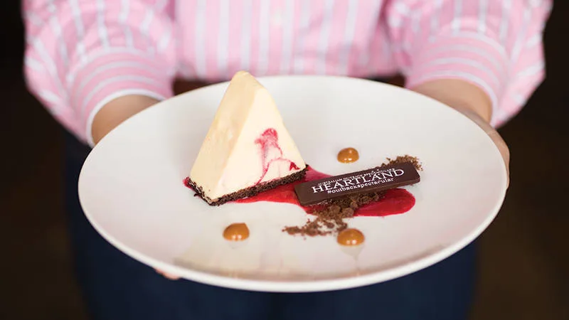 A person in a pink striped shirt holds a white plate with a triangular cheesecake, raspberry sauce, chocolate garnish, caramel dots, and a piece labeled "HEARTLAND.