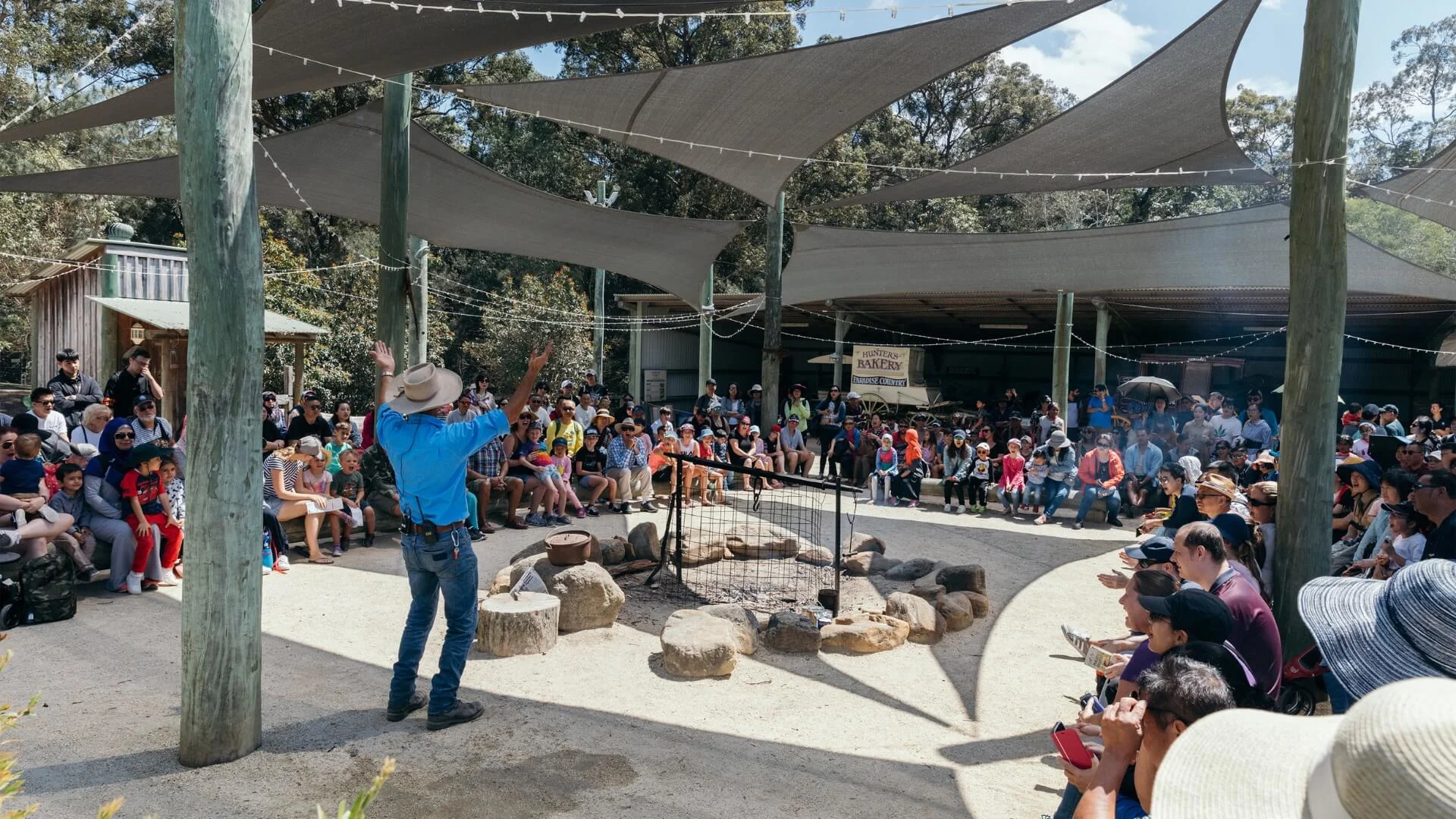 Crowd gathered at Paradise Country watching an outdoor Australian farm show with a presenter in a cowboy hat demonstrating by a campfire setup.