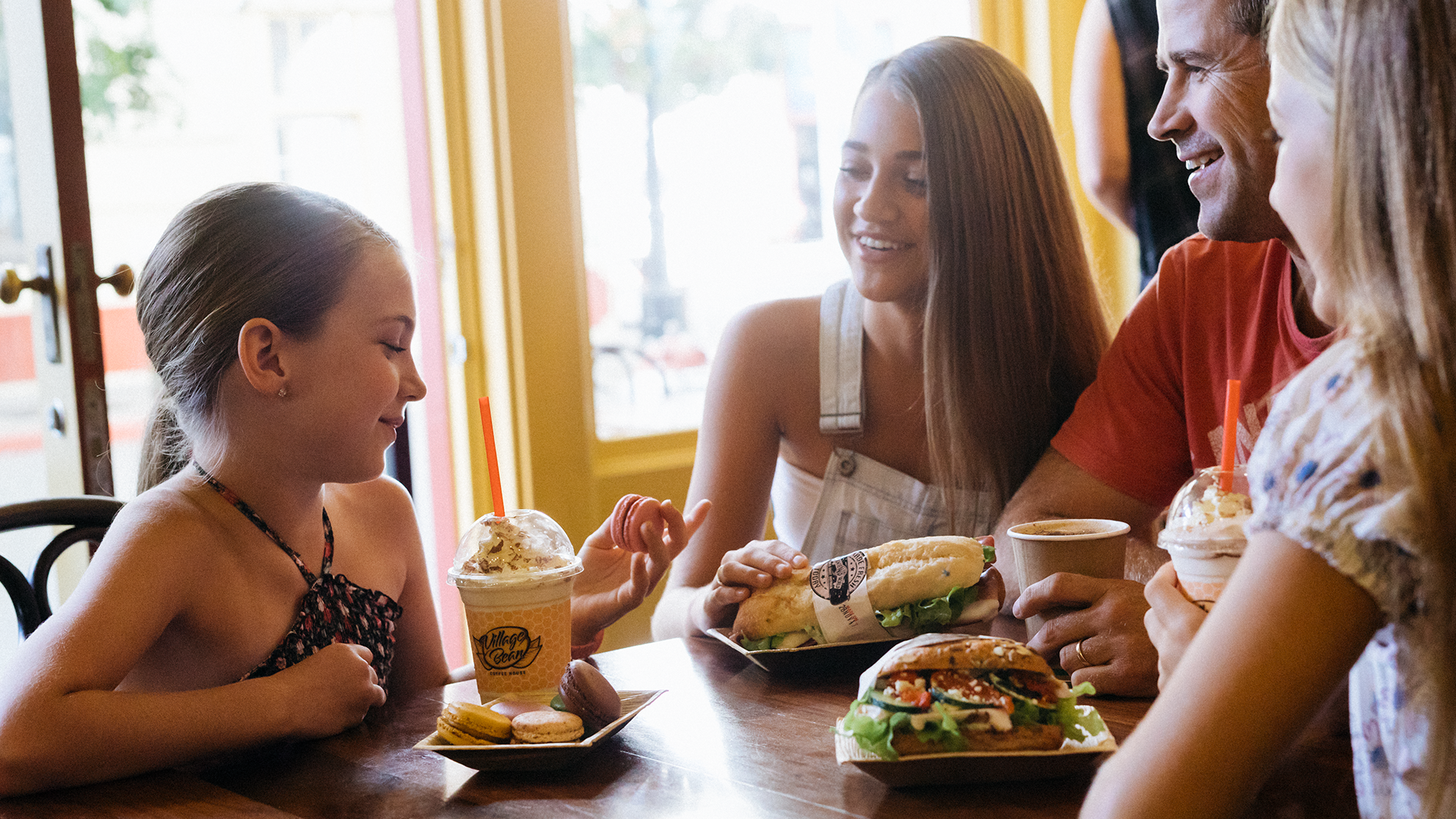 A family sitting at a table, happily enjoying food and drinks at Village Bean at Warner Bros. Movie World, with a cozy and inviting atmosphere. foodandbeverage
