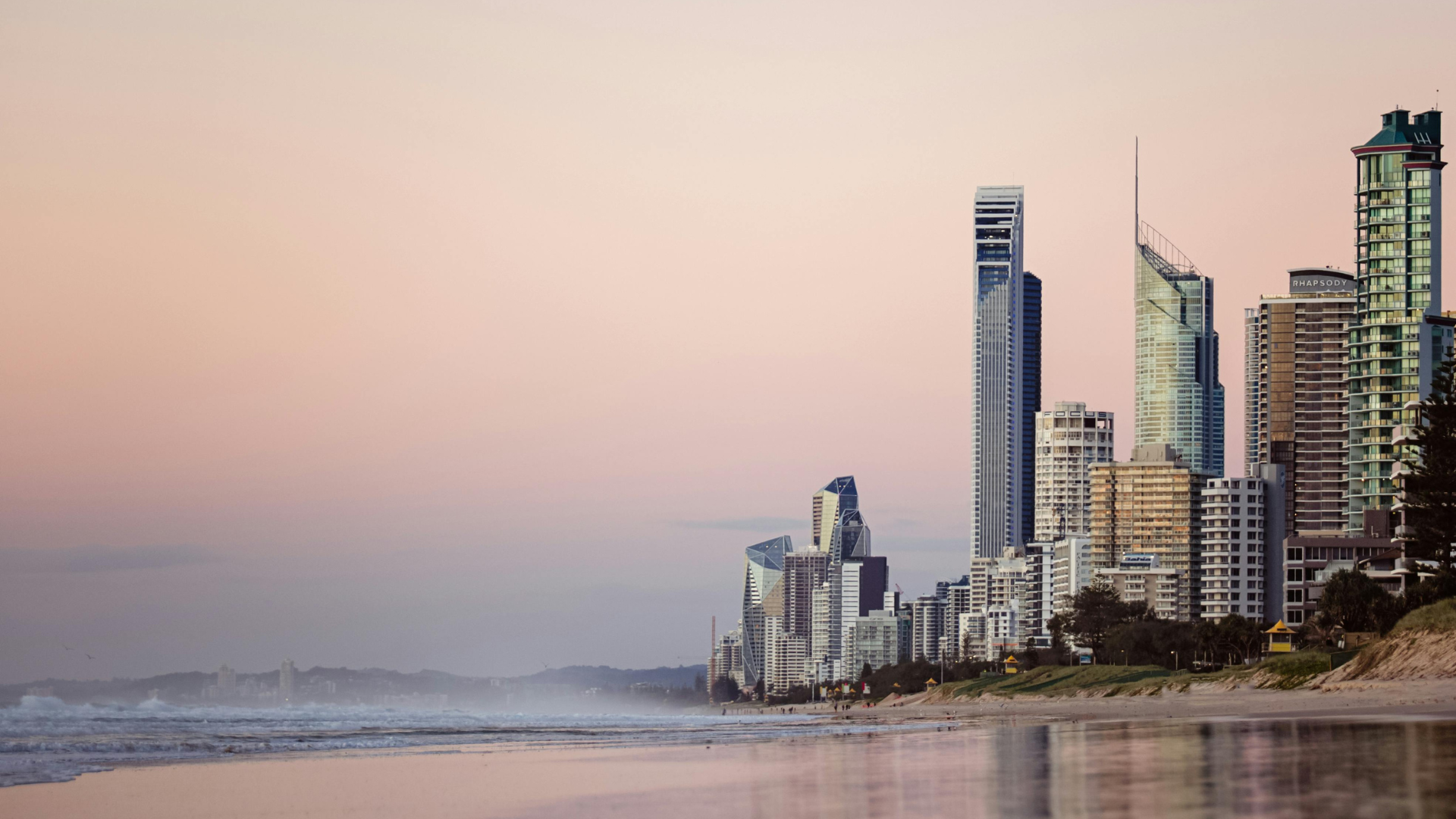 Skyscrapers and modern buildings line a sandy beach at sunrise or sunset, with a calm ocean and soft pastel colors in the sky, creating a peaceful coastal cityscape.