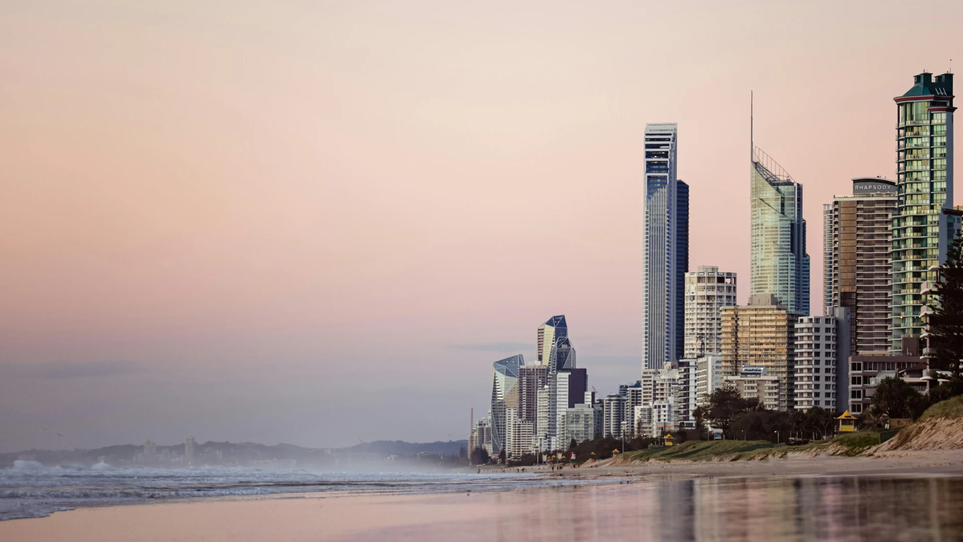 Skyscrapers and modern buildings line a sandy beach at sunrise or sunset, with a calm ocean and soft pastel colors in the sky, creating a peaceful coastal cityscape.