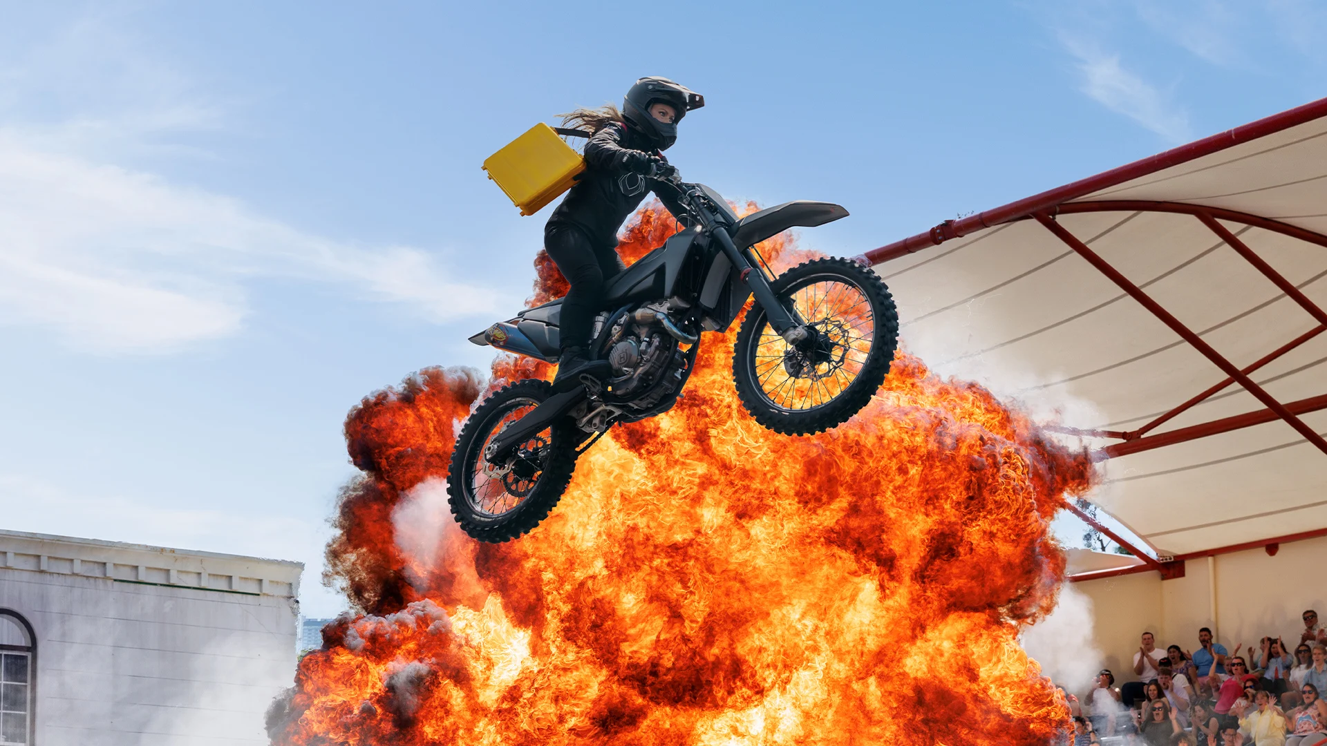 A person wearing a helmet jumps a motorcycle over a large explosion during a stunt show, with spectators watching from the stands in the background.