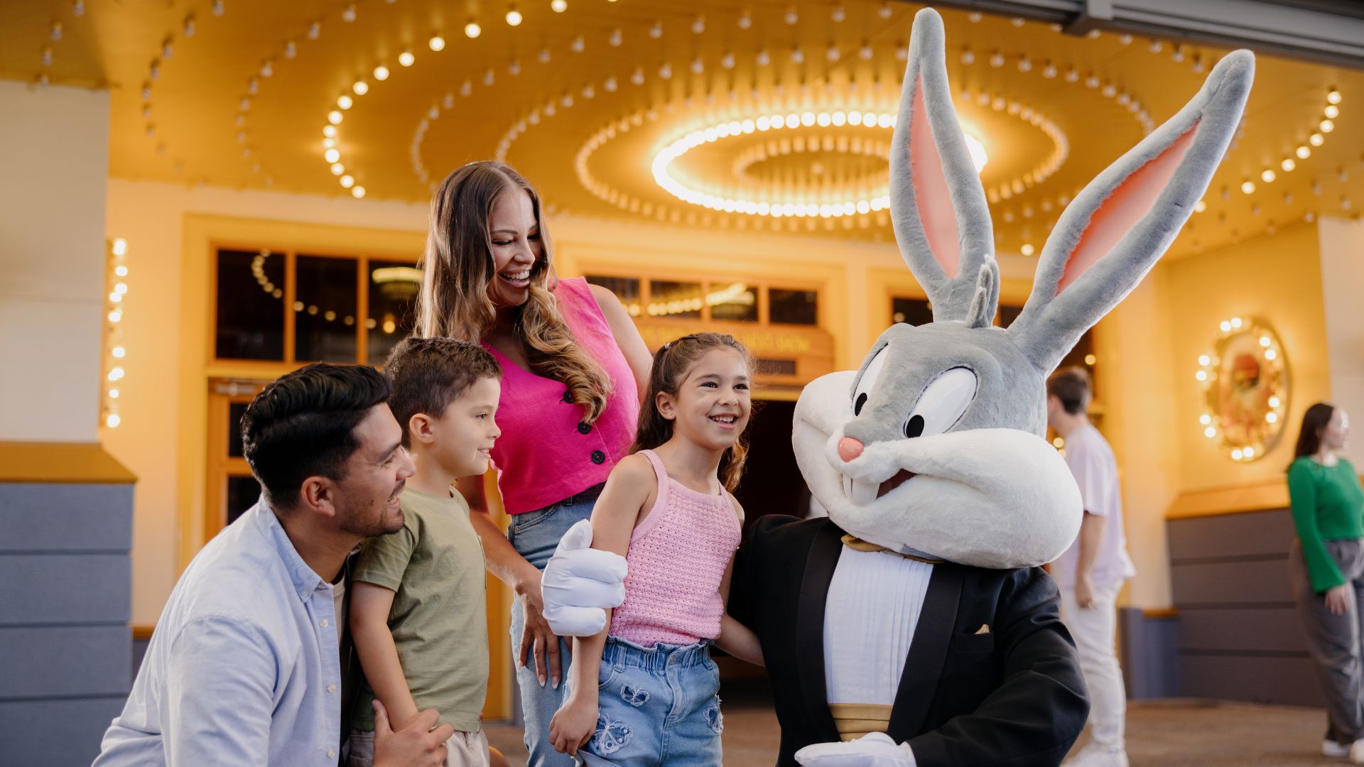 A smiling family of four poses with a person in a Bugs Bunny costume outside a brightly lit building. The parents and two children look happy and excited.
